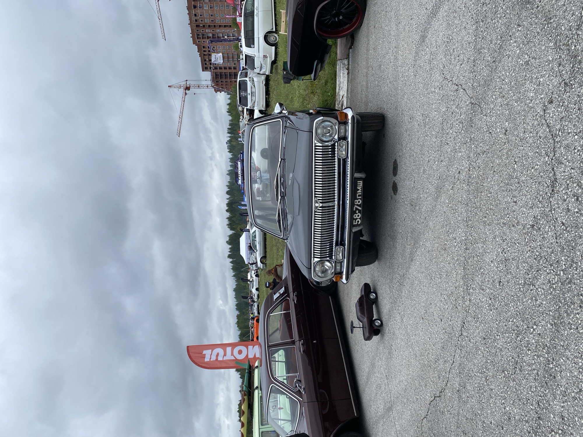 This image captures an outdoor car show, possibly at the Hippodrome in Perm, Russia, under a cloudy sky. The foreground is dominated by a black classic sedan with a license plate displaying "58-78 ПМШ." To its left, a maroon classic sedan is partially visible, and a small toy car sits on the asphalt between them. In the background, other vehicles, including white vans and sedans, are parked on grass and asphalt. Construction cranes and buildings are visible in the far distance. A red banner with the word "MOTUL" stands behind the maroon car. The ground is a cracked asphalt surface.