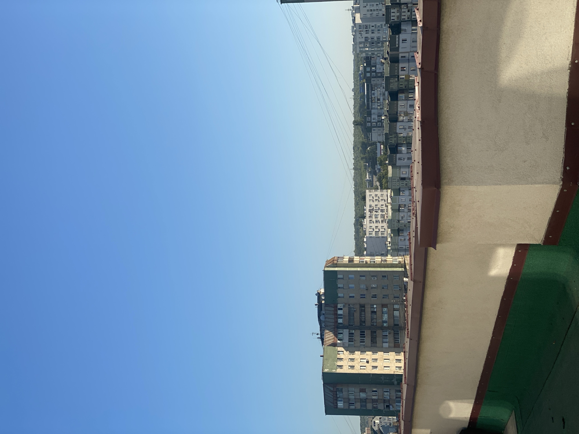 The image depicts a view of a city skyline under a clear blue sky, taken from an elevated vantage point, possibly a rooftop. In the foreground, there is a light-colored parapet wall with a reddish-brown trim and a green surface below, suggesting a balcony or rooftop area. Beyond the parapet, a dense urban landscape unfolds, characterized by numerous apartment buildings of varying heights and architectural styles. One prominent building in the mid-ground is a multi-story apartment block with a light beige and green facade, featuring many windows. Further in the distance, rows of similar apartment buildings stretch across the horizon, interspersed with patches of greenery and trees, indicative of a residential district. Overhead, thin power lines crisscross the sky, adding to the urban infrastructure. The scene suggests a typical residential neighborhood in the city of Chișinău, Moldova, likely during daytime due to the bright sky and shadows. There are no people or animals visible in the image, and no specific actions or interactions are captured.