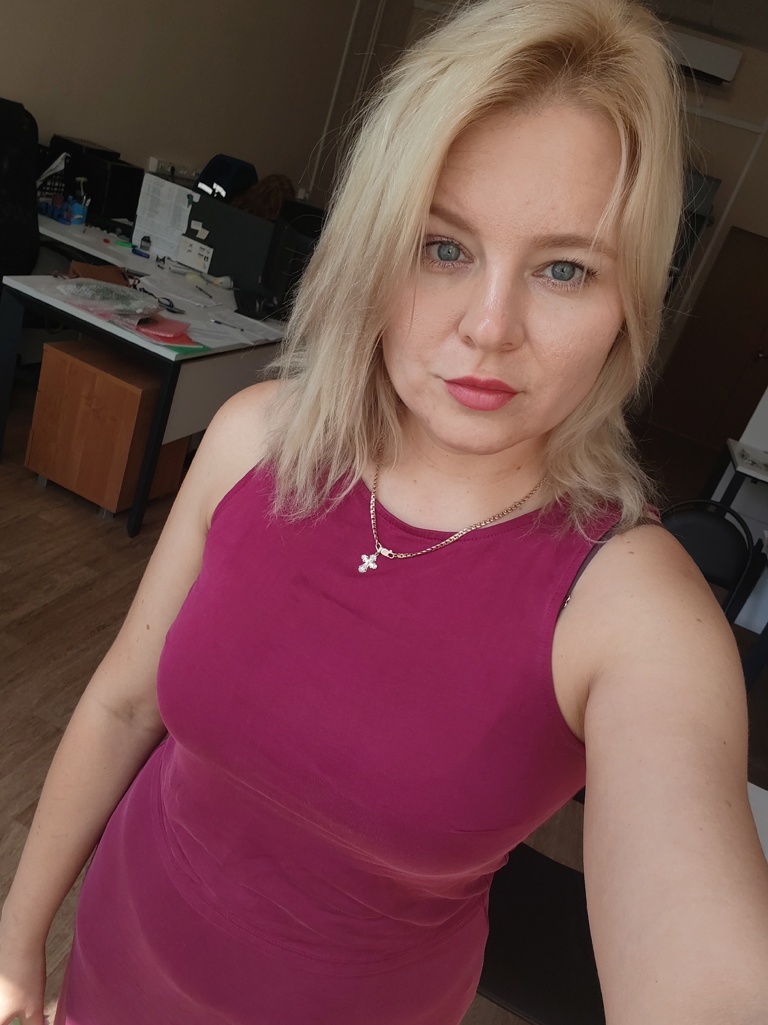 The image shows a selfie of a blonde woman with blue eyes and pink lipstick. She is wearing a sleeveless magenta dress and a gold necklace with a cross pendant. She appears to be in an office setting, with desks, computers, and office supplies visible in the background. The overall impression is that of a casual office selfie.