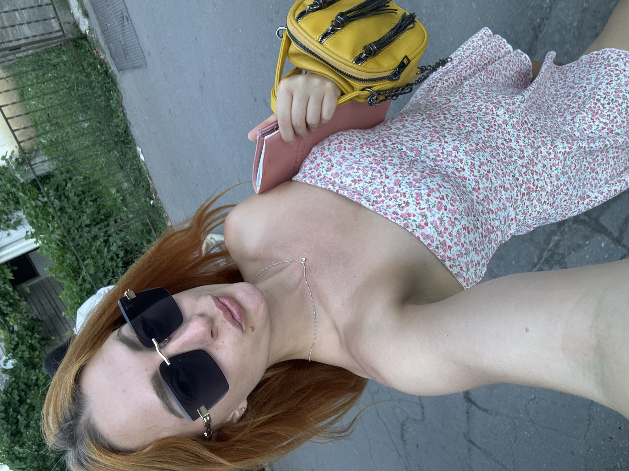 A young woman with auburn hair is standing on a paved street, likely in Budva, Montenegro, given the presence of lush green foliage and a weathered metal fence. She is wearing a strapless, floral-print dress and large, dark, rimless sunglasses. In her right hand, she holds a pink notebook or folder, and to her right, a small yellow purse with a chain strap is visible. Her left arm is extended, suggesting she is taking a selfie. The background features a building and a drain grate on the pavement.