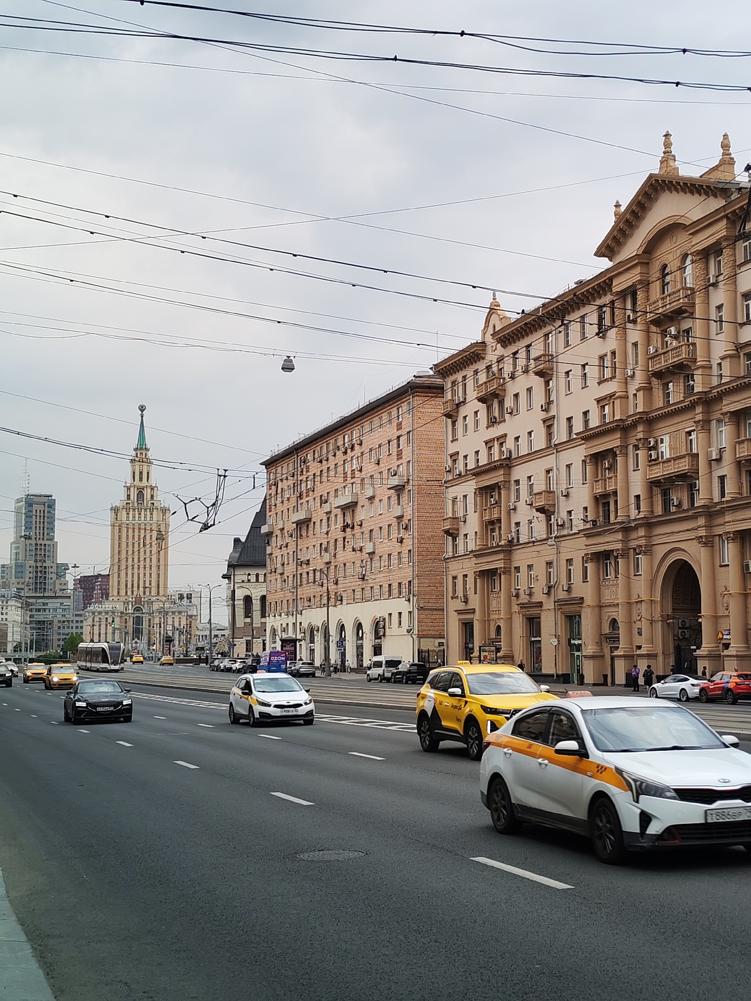This is a street-level, eye-level, wide shot of a busy city street in Moscow, Russia, under an overcast sky. The scene captures a section of a wide avenue with multiple lanes of traffic. Several vehicles are visible, including yellow taxis, a white sedan with yellow accents that appears to be a taxi, a black sedan, a yellow SUV, a tram, and a white van. The buildings lining the street are large, multi-story structures with ornate architecture, typical of Stalinist-era buildings in Moscow. One prominent building on the left is a tall, art deco skyscraper with a spire. On the right, a long, tan building features a grand facade with columns and balconies. The street is lined with sidewalks where a few pedestrians are visible. Overhead, a complex network of power lines and tram wires crisscrosses the sky. The overall impression is of a bustling urban environment during daytime.
