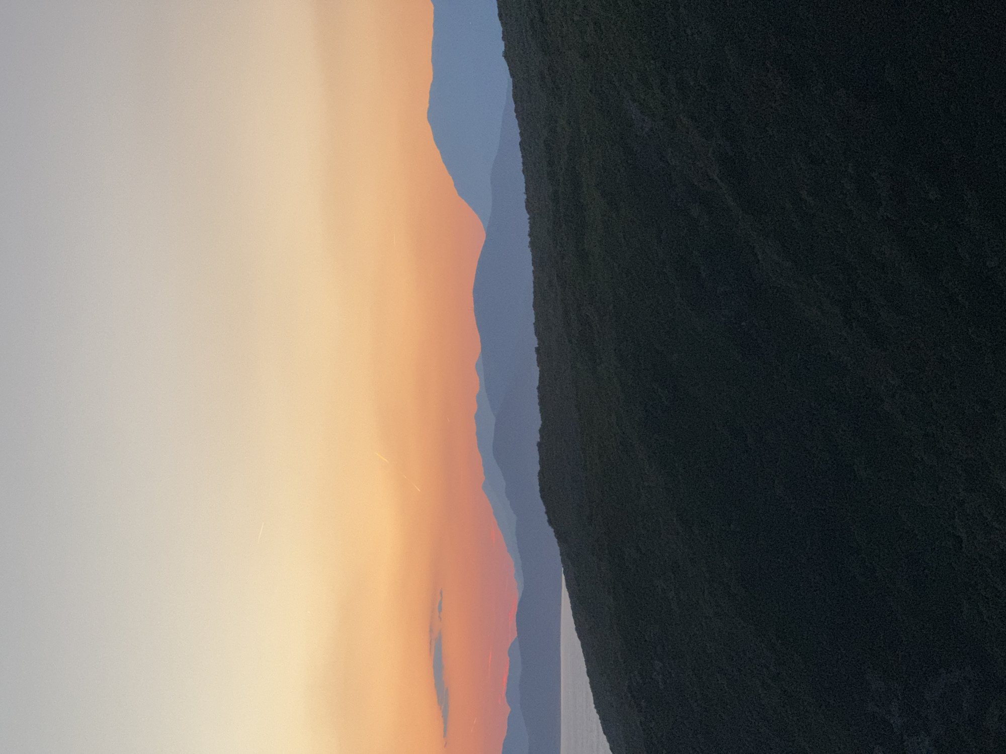 The image depicts a serene sunset over a mountainous coastal landscape, likely in Glavatičići, Montenegro. The sky transitions from a soft pale blue at the top to a warm gradient of orange and pink hues near the horizon. Silhouetted against the colorful sky are several layers of mountains, receding into the distance. In the foreground, a dark, densely vegetated hillside slopes downwards towards a body of water, possibly the Adriatic Sea, which is visible on the left side of the frame. The overall scene is tranquil and natural, characterized by its natural beauty and subdued lighting. There are no people or man-made objects visible in the frame, emphasizing the unspoiled, natural environment.