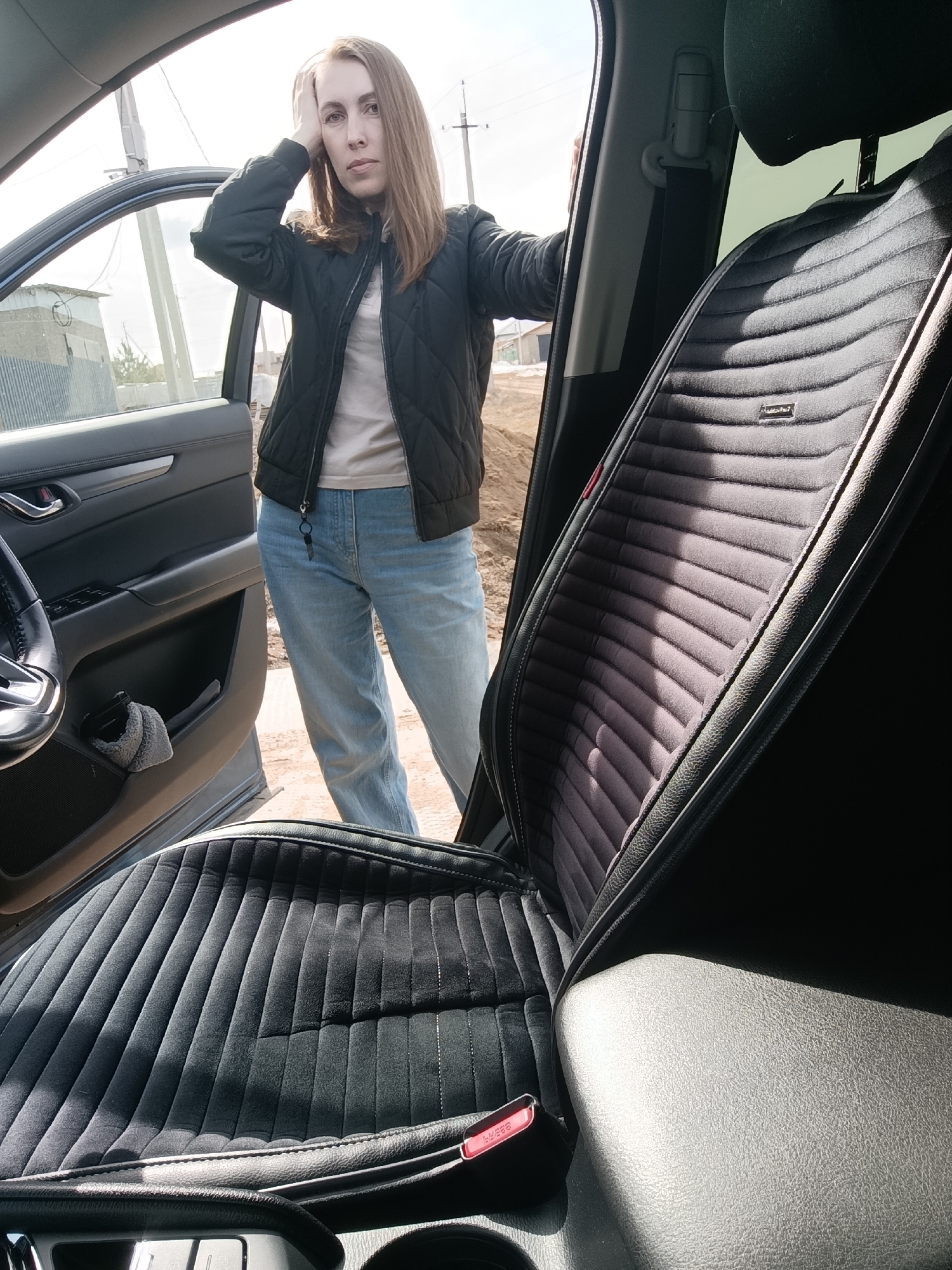 A woman stands outside an open car door. She wears a dark quilted jacket, light-colored t-shirt, and light blue jeans. Her left hand is on her head, and her right hand rests on the car door frame. The car seat is visible, covered in a dark, ribbed fabric. Outside the car, the ground appears to be dirt and gravel. A building and power lines are visible in the background. The location is Chaikovsky, Russia.