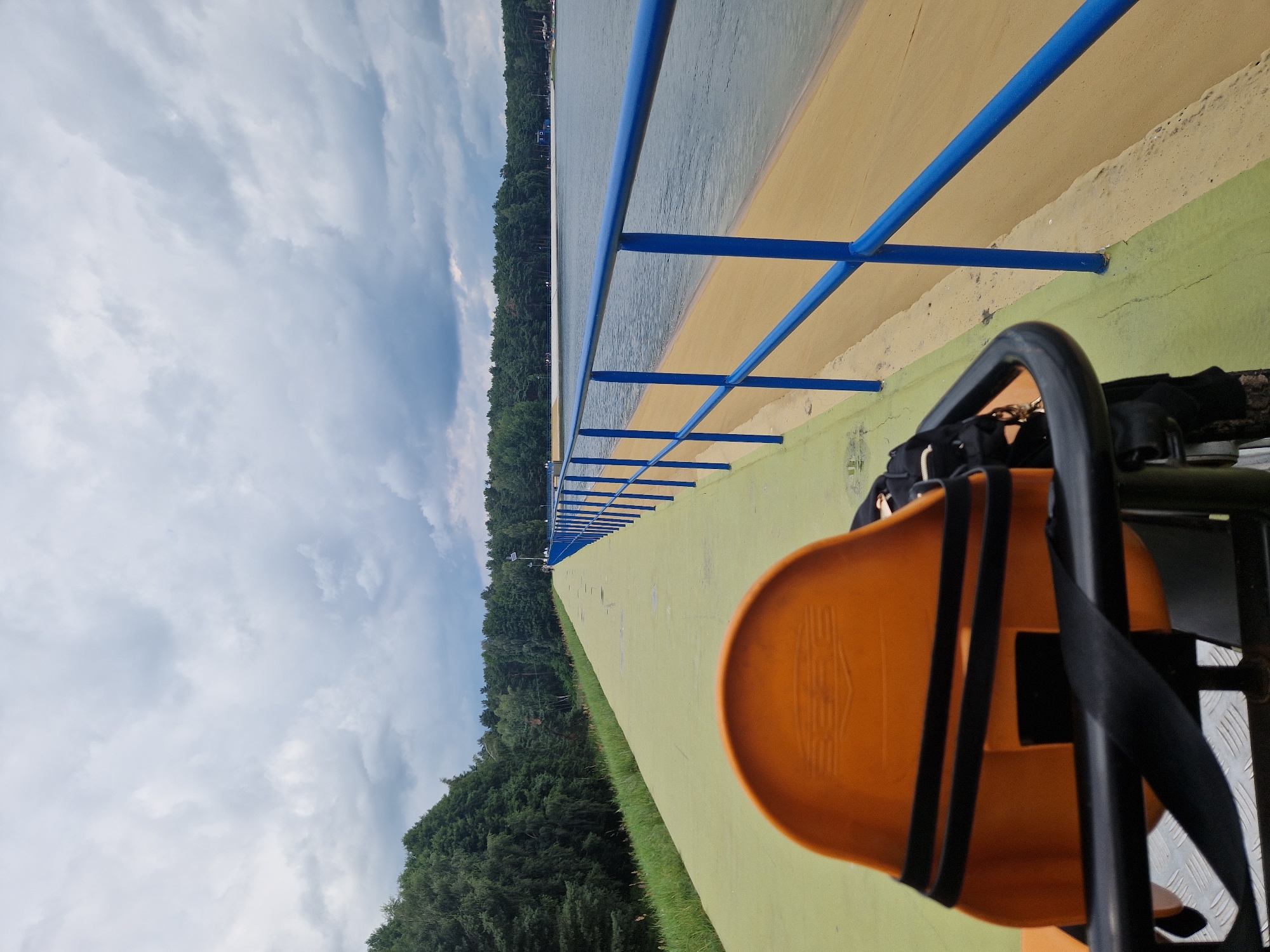 The image shows a person's perspective from a vehicle or a seating area, looking down a paved pathway next to a body of water. In the foreground, an orange seat with black straps is visible, part of the vehicle or seating arrangement. The pathway is painted green and stretches into the distance, bordered by a sandy area and a blue metal railing. Beyond the railing is a large body of water. The background features a dense line of green trees, and the sky is overcast with gray clouds. There are a few distant figures walking on the pathway and some boats visible on the water. The overall scene depicts an outdoor recreational area, possibly a dam or a waterfront promenade.