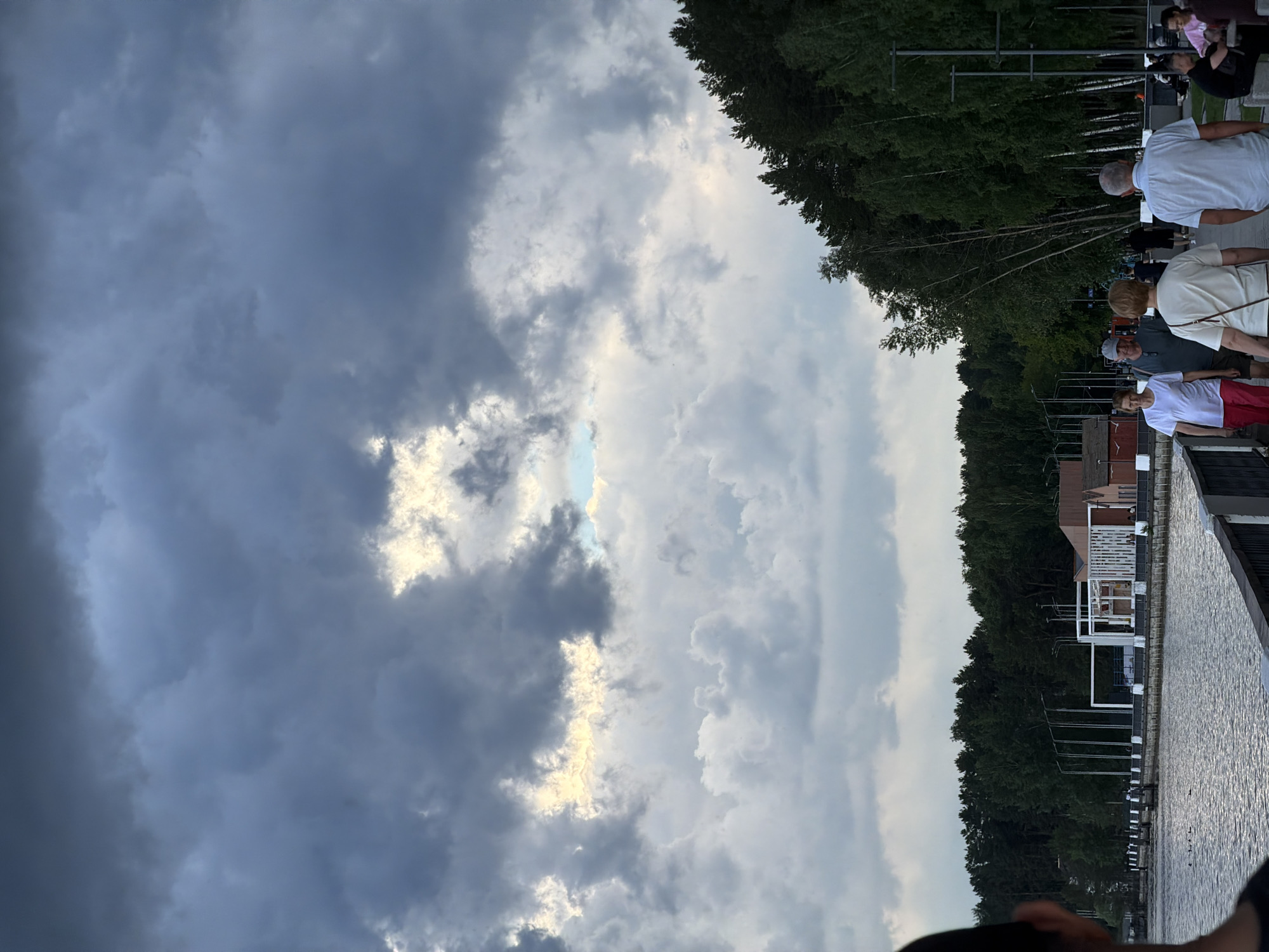 The image depicts a waterfront promenade in the city of Chaikovsky, Russia, under a dramatic sky filled with heavy, dark clouds. A break in the clouds reveals a patch of bright sunlight. On the right side of the image, a dense line of trees, primarily birch and pine, flanks the promenade. Several people are walking along the promenade, some in small groups and others individually. One group in the foreground consists of a woman in a white t-shirt and pink pants, a man in a grey t-shirt and baseball cap, and another woman in a cream-colored top. Further down the promenade, other individuals and small groups are visible, some sitting on benches. The water body, likely the Kama River, is calm with ripples. Along the waterfront, there are modern architectural structures, including white geometric frameworks and low-rise buildings, suggesting a developed recreational area. Streetlights are also visible along the promenade. The overall atmosphere is overcast, hinting at approaching rain.