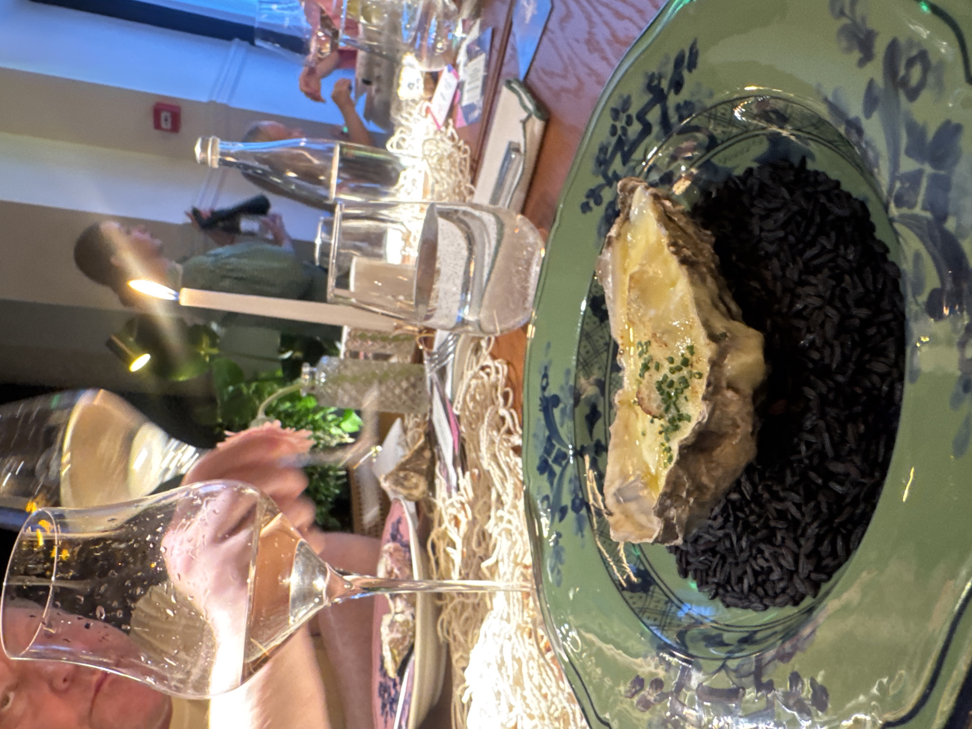A close-up, eye-level shot shows a single baked oyster served on a bed of black rice on a green, patterned plate. The oyster is topped with melted cheese and finely chopped chives. In the background, blurred figures and table settings suggest a restaurant environment. A tall, lit candle is visible to the left of a glass of water. The lighting is dim and warm.