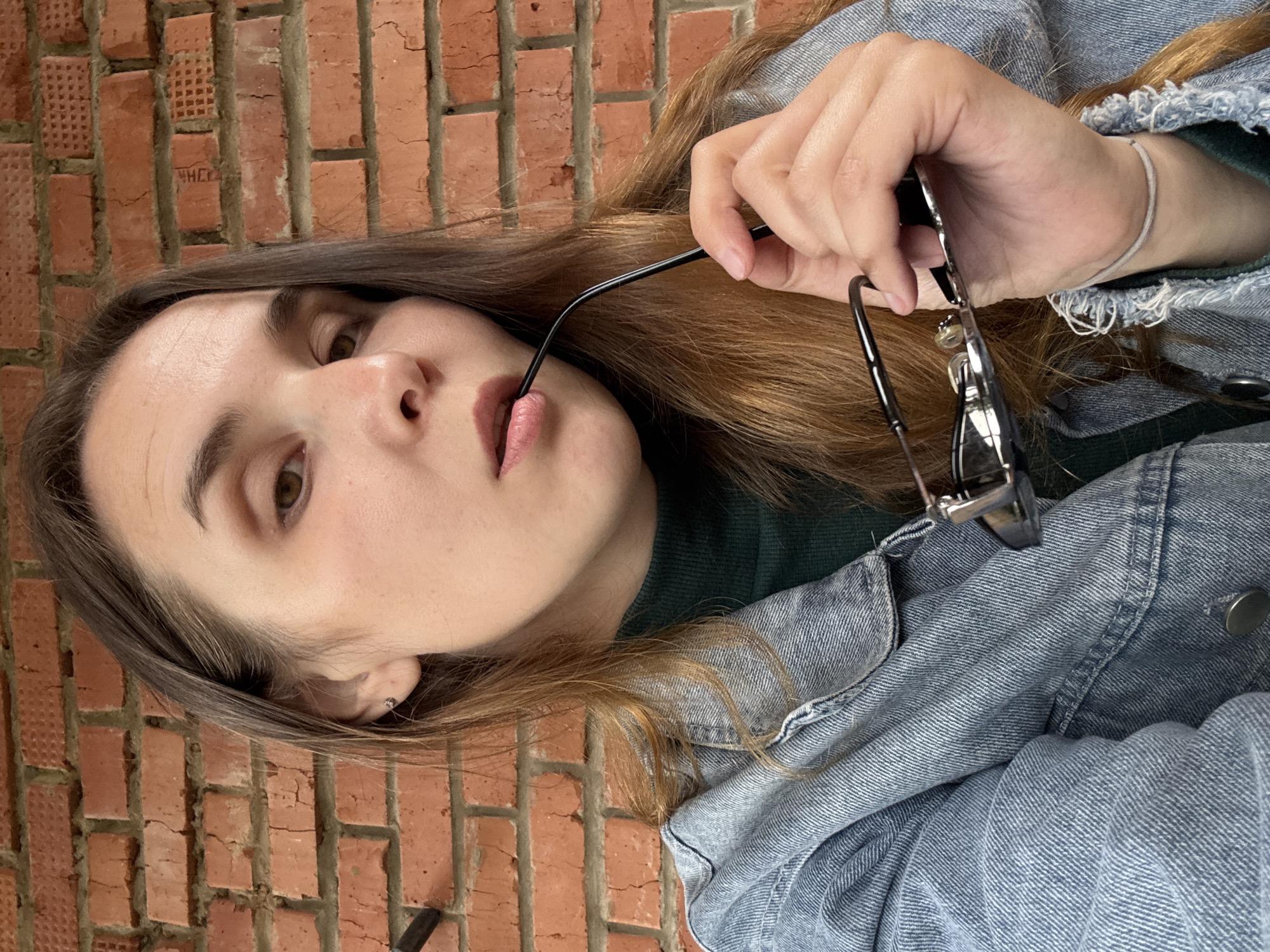 A medium-shot, chest-up, selfie-style photograph features a young Caucasian woman with long, light brown hair and light brown eyes. She is wearing a dark green turtleneck sweater and a light blue denim jacket. The woman is holding a pair of black-rimmed glasses in her right hand, with the earpiece lightly touching her lips. She is looking directly at the camera with a neutral expression. The background consists of a textured, reddish-brown brick wall. The lighting suggests an outdoor or well-lit indoor environment. There is no clear indication of the specific location within Votkinsk, Russia.