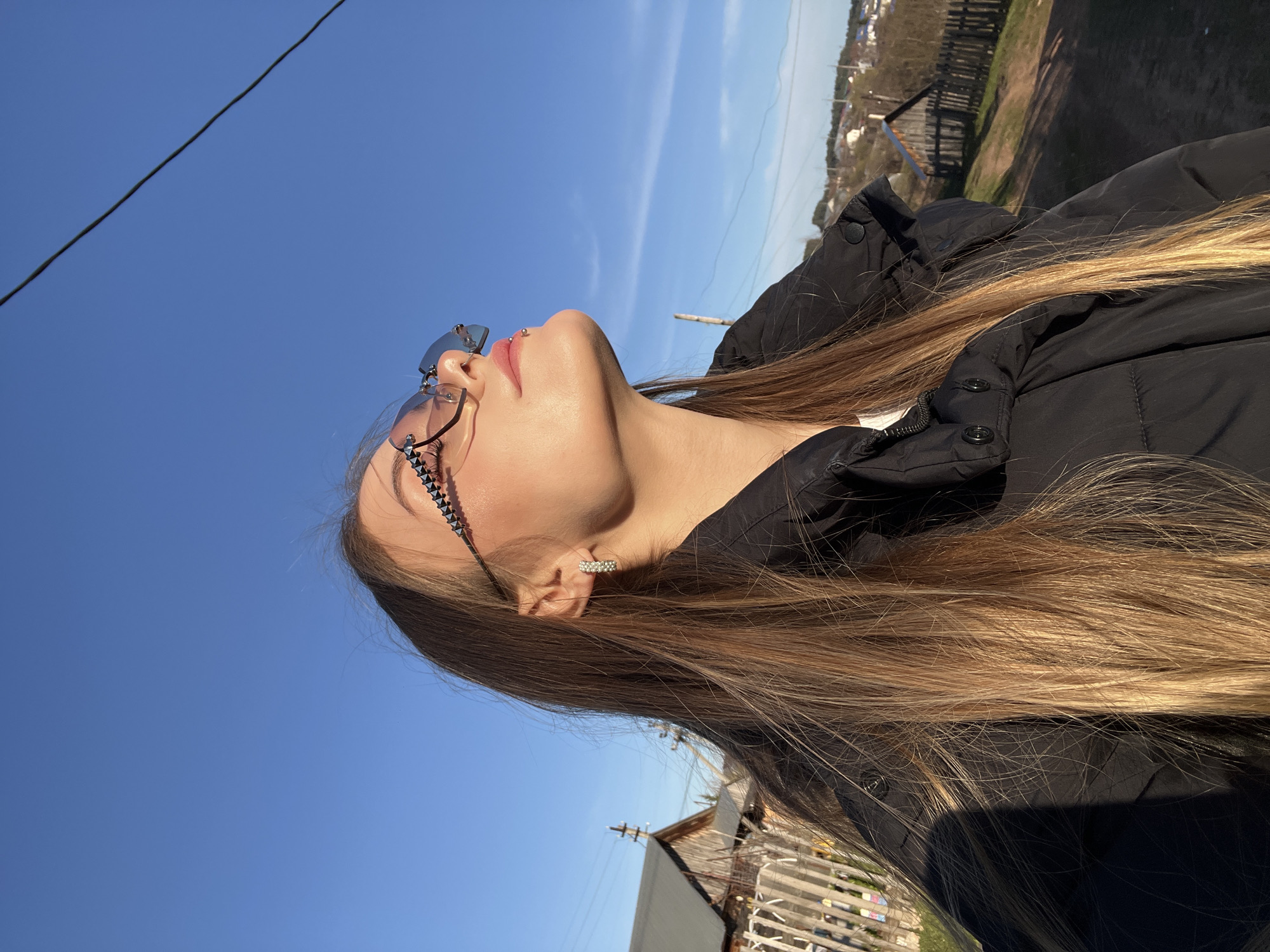 A woman with long blond hair and sunglasses is looking up at a blue sky. She is wearing a black jacket and has an earring in her left ear. In the background, there is a building with a wooden fence. The image was taken in Фоки, Russia.