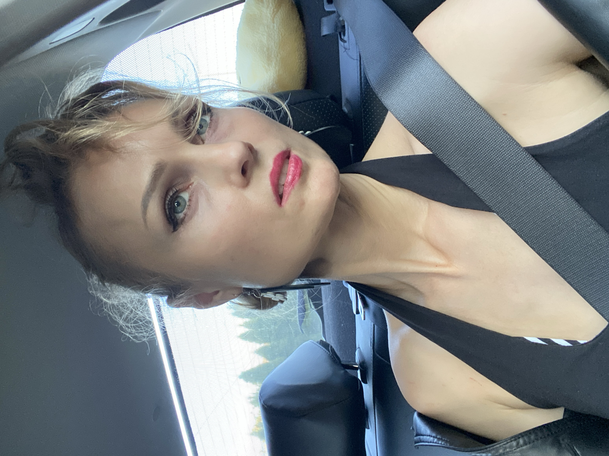 A medium shot shows a woman in a car, possibly in Lida, Belarus. She is looking towards the camera with a slight smirk and her eyes are rimmed with black eyeliner. Her lips are painted a bright pink. Her hair is pulled up in a messy bun with strands falling around her face. She is wearing a black halter dress and a seatbelt is fastened across her chest. In the background, there are car seats and a window with a blurred view of trees. A fluffy yellow pillow is visible on the passenger seat.