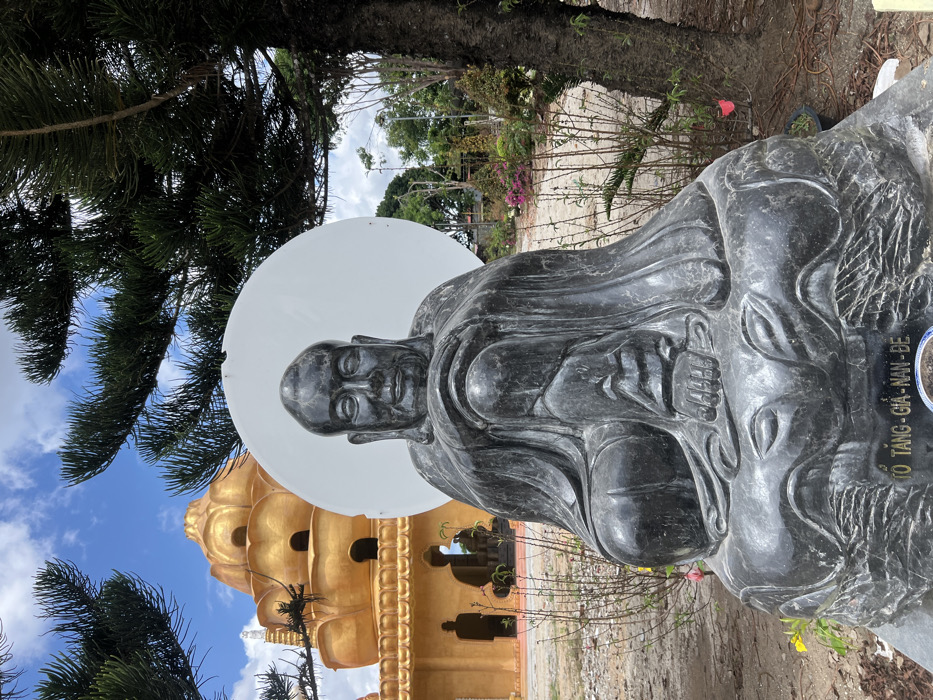 A dark grey stone statue of a seated figure, identified by the inscription "TỔ TĂNG - GIA - NAN - ĐỀ" on its base, occupies the central foreground of an outdoor scene in Đăk Mar, Vietnam. The figure is bald, possesses a serene facial expression, and is draped in robes with hands resting in its lap. A large, plain white circular disk is positioned behind its head, creating a halo effect. In the midground to the left, a prominent multi-tiered, golden architectural structure, resembling a pagoda or temple with lotus-petal-shaped roofs, is visible. Dark silhouettes of additional statues are discernible through the archways on the ground level of this golden building. The surrounding ground consists of exposed reddish-brown soil with sparse, low-lying, bare-branched vegetation and a few potted plants. A large conifer tree trunk is present on the far right, with its dark green branches extending into the upper right and left portions of the frame. The sky is blue with scattered white clouds.