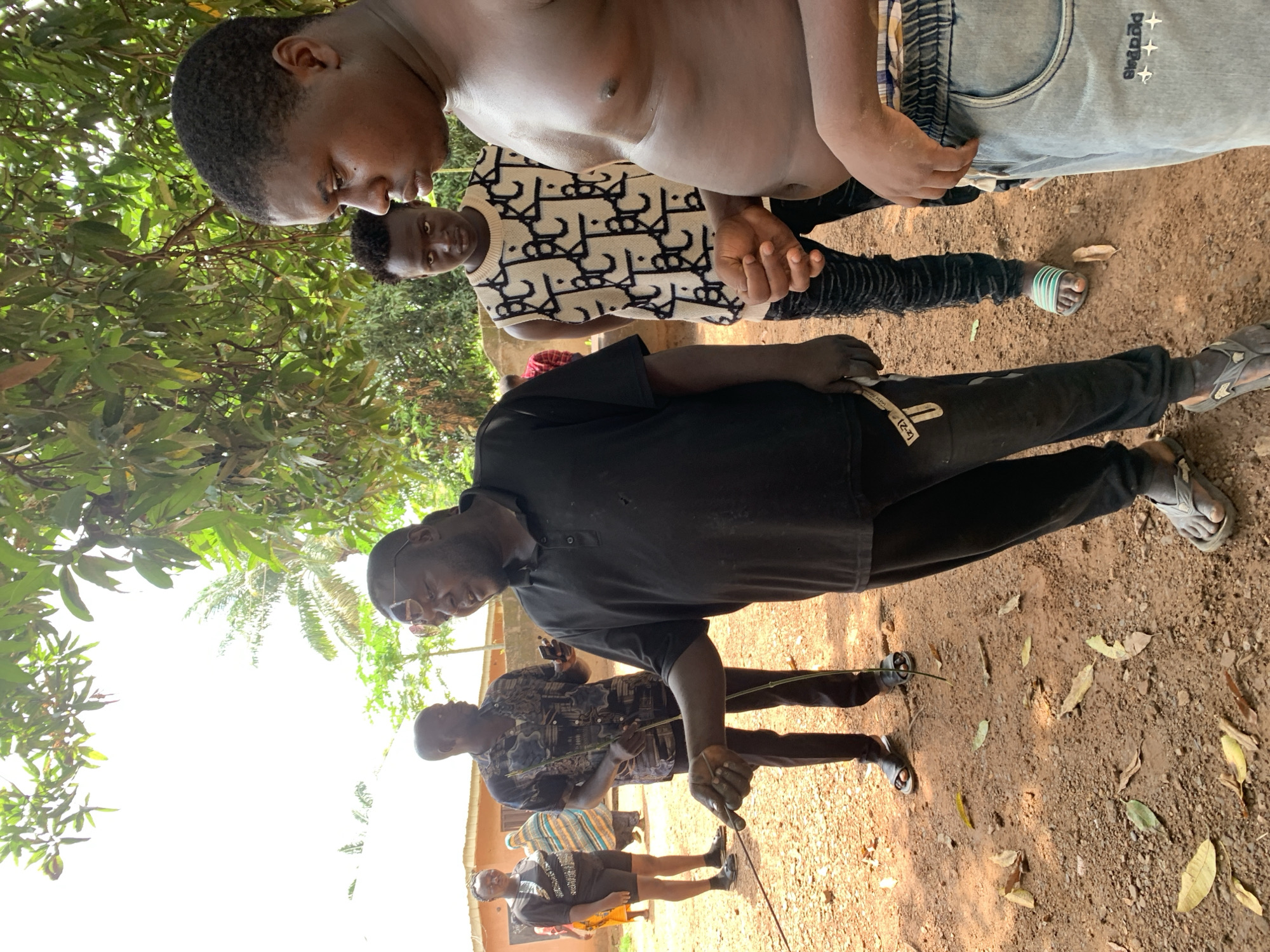 Four individuals, three males and one female, are standing on a dirt ground outdoors. The male in the foreground, who appears to be middle-aged, is wearing a black polo shirt and dark pants. He is gesturing with his right hand towards the ground, holding a thin stick. To his left, a younger male in a white sleeveless shirt with a black pattern and black pants stands looking in the same direction. Behind them, another male in a patterned shirt and dark pants is holding a stick and a phone. In the background, a female in a black t-shirt and shorts is visible. The environment suggests a rural or semi-urban setting in Akwanga, Nigeria, with a building and trees in the background. The ground is covered in reddish-brown dirt with scattered leaves.