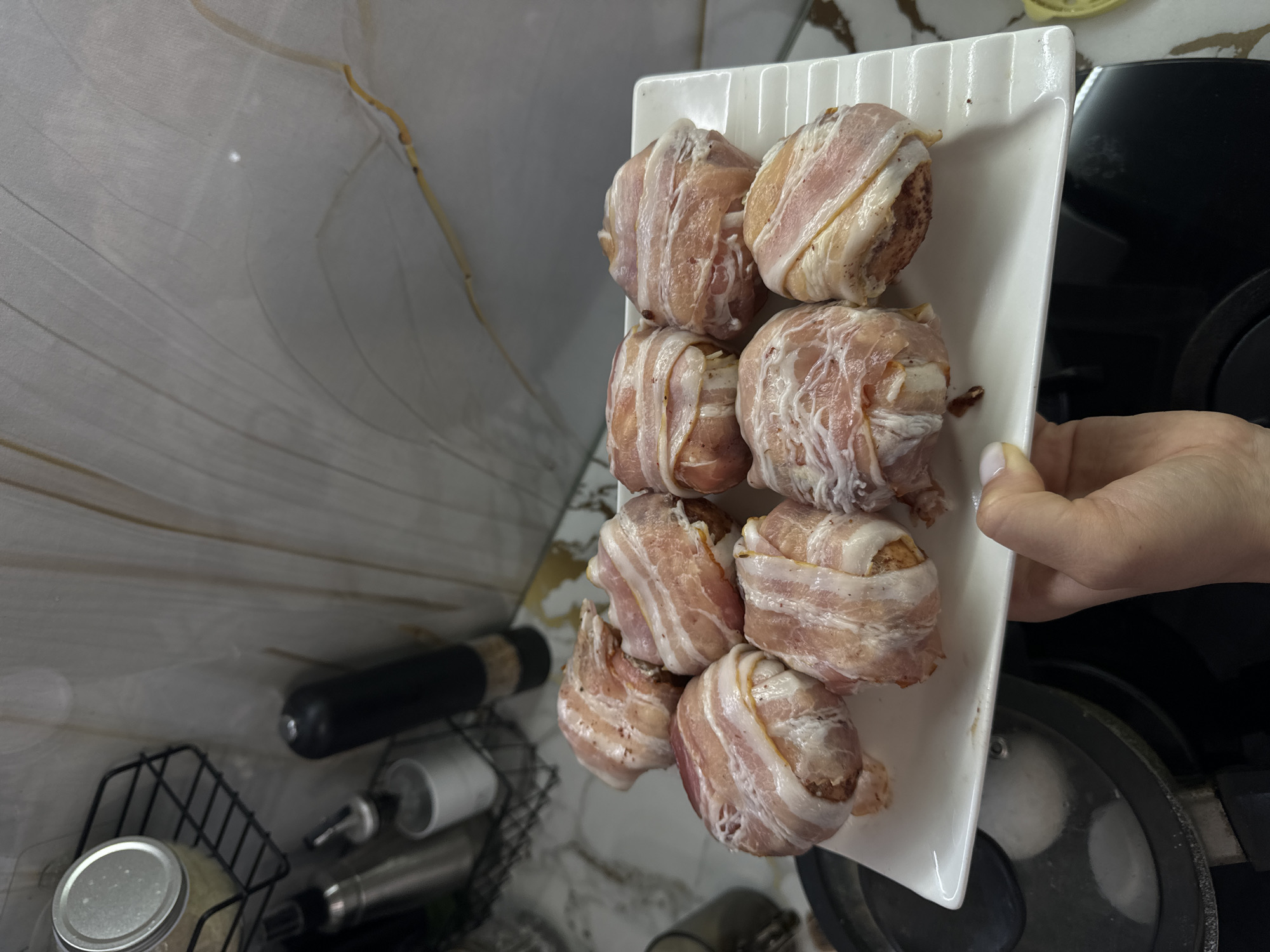 In a kitchen environment, a white rectangular plate holds eight food items, each wrapped in bacon. The food items appear to be meatballs or similar spherical food. The plate is held by a person whose hand is visible in the lower right portion of the frame. In the background, a tiled wall and a shelf with various kitchen items are visible. The scene is set in a domestic kitchen, possibly in Поназырево, Russia.