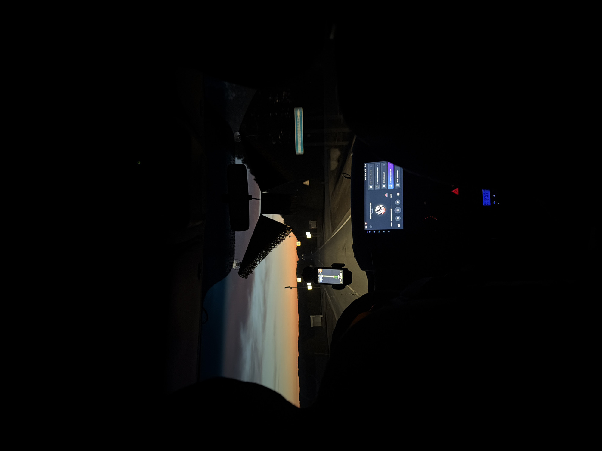 A nighttime view from inside a car traveling along a road. Through the windshield, a sunset sky is visible with colors transitioning from orange to blue. Roadside lights illuminate the roadway. Inside the car, a navigation device is mounted on the dashboard, and a music player is displayed on a large screen. The scene appears to be in Kirillovka, Russia based on the sign at the top of the windshield.