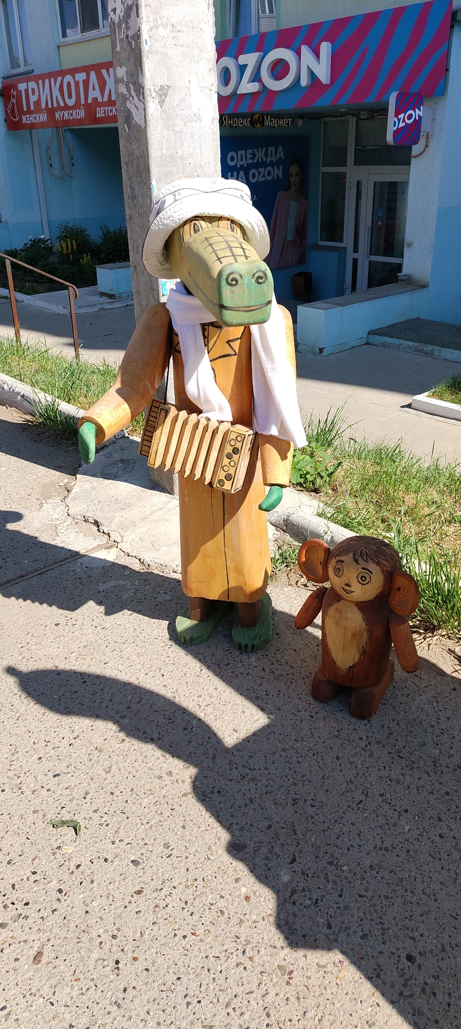 The image depicts a sunny outdoor scene in front of an OZON store in Chaikovsky, Russia. Two wooden figurines are prominently featured: a tall alligator wearing a white hat, scarf, and holding an accordion, and a smaller, brown, bear-like character with large ears. The alligator figurine stands on a cracked concrete sidewalk, casting a long shadow. The smaller figurine is positioned to the right of the alligator, also on the sidewalk. In the background, the OZON store's facade is visible with its characteristic blue and pink striped signage. A smaller sign reads "Яндекс Маркет" (Yandex Market) and another displays "ОДЕЖДА НА OZON" (Clothing on OZON). To the left, a sign for "ТРИКОТАЖ" (Knitwear) is visible, indicating a clothing store. The environment appears to be a commercial area with a sidewalk, grass, and a building.