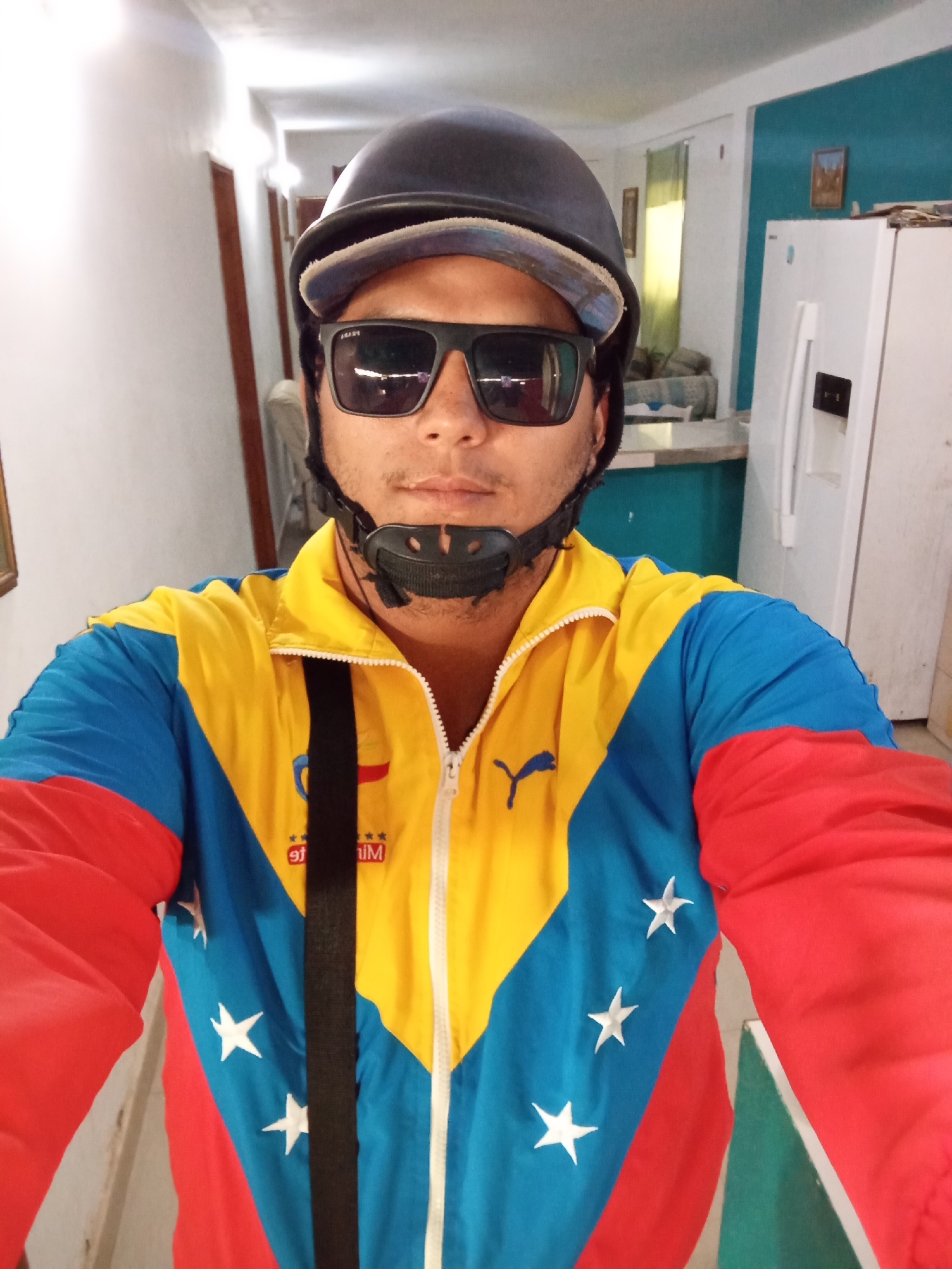 A man wearing a helmet and sunglasses stands indoors. He is wearing a zip-up jacket with the colors of the Venezuelan flag (yellow, blue, and red) and white stars. A black strap is draped across his chest. Background shows an interior with white walls and a white refrigerator. Location is Juan Griego, Venezuela.