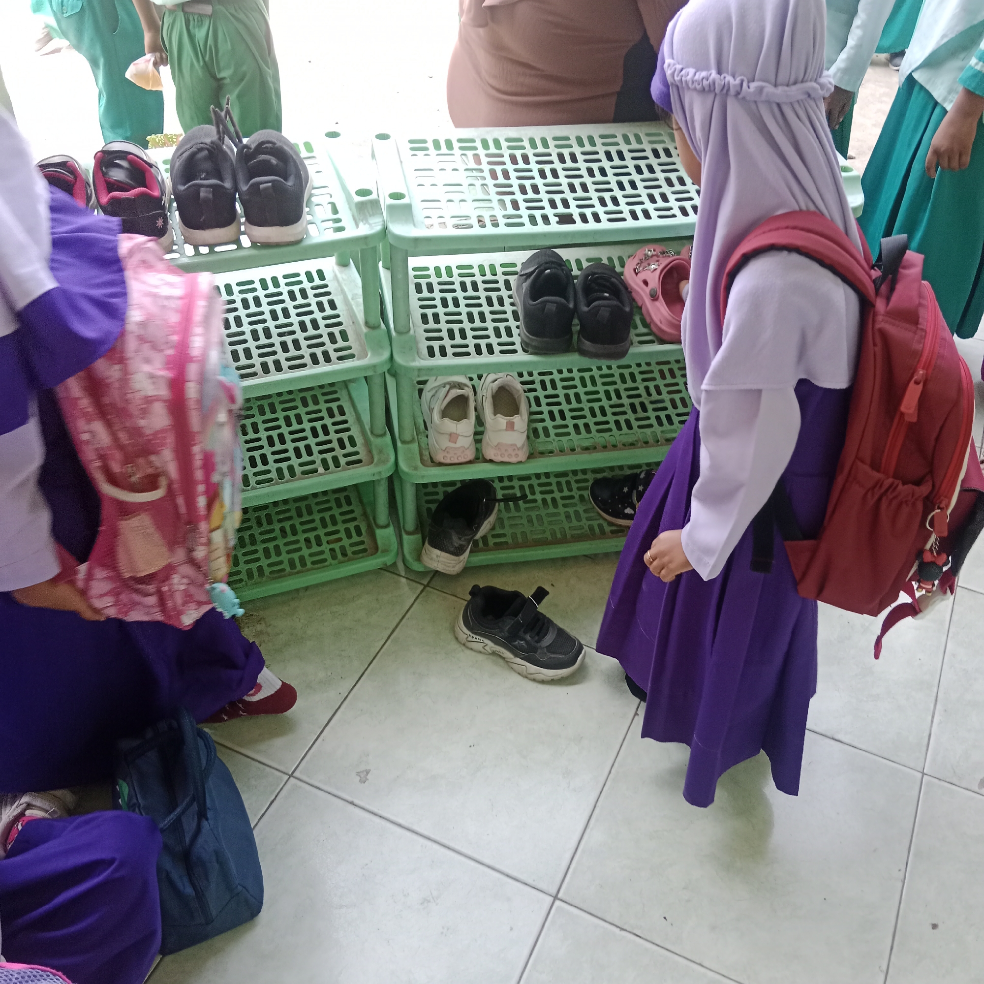 A group of young individuals, likely students, are gathered near a multi-tiered green plastic shoe rack. The shoe rack is filled with various shoes, including black sneakers, pink sandals, and white sneakers. Some of the individuals are wearing school uniforms, characterized by purple and white or green garments. One individual in the foreground is wearing a purple hijab and a purple skirt, with a red backpack slung over their shoulder. Another individual in the background is wearing a green uniform. The floor is tiled with light-colored square tiles. The scene appears to be an outdoor or semi-outdoor area, possibly a school yard or entrance, given the presence of shoe racks. The location is specified as Tegowanu Kulon, Indonesia. The time of day is indeterminate, but the lighting suggests daytime.
