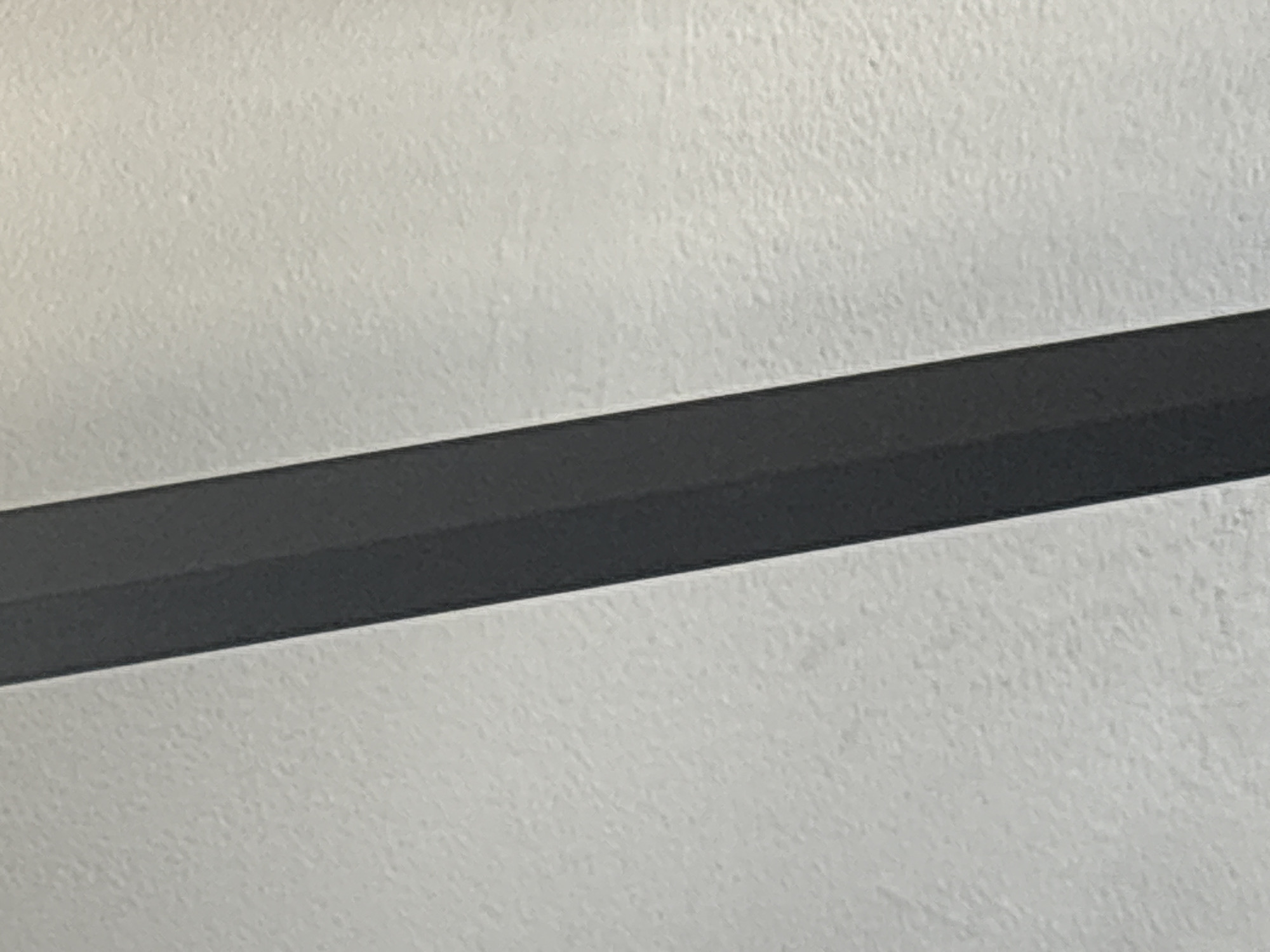The media file shows a close-up, angled shot of a dark, beveled bar against a textured, light-colored background. The bar is positioned diagonally across the frame, running from the bottom left to the top right. It has a matte finish and appears to be made of a solid material. The background has a rough, somewhat uneven texture, suggestive of a wall or a textured surface. The lighting appears even, with no distinct shadows that would indicate a specific time of day or weather conditions. There is no discernable activity or event taking place, and no people are visible. The image is purely an abstract composition focusing on the geometric form of the bar and the texture of the background. There is no visible text in the image.