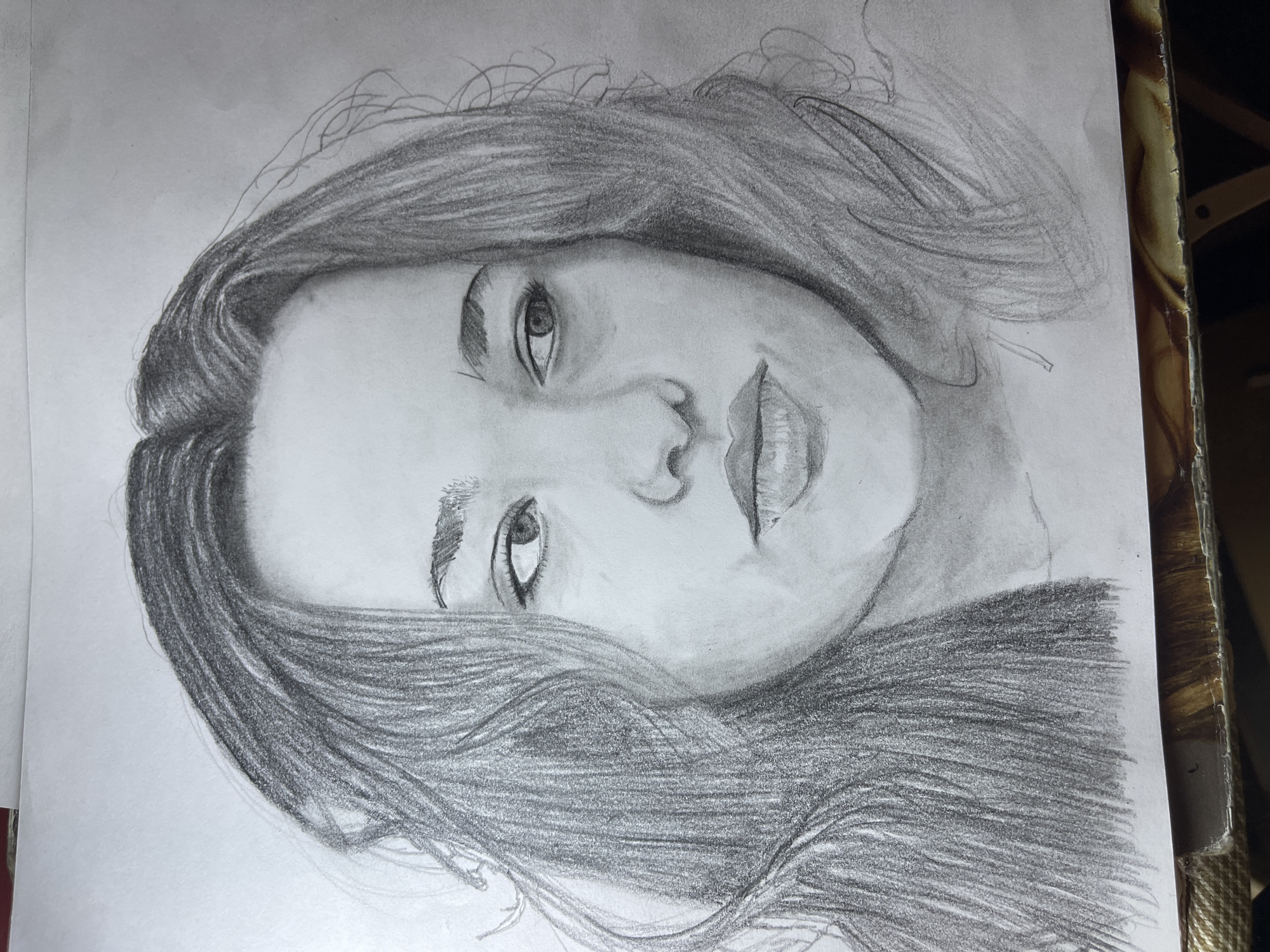 A pencil drawing of a woman's face and hair. The face is angled, slightly rotated. The woman has long, dark hair. The drawing is on a piece of white paper. The background is blurred and indistinct. The scene is not in Jos, Nigeria; it appears to be on a table indoors.