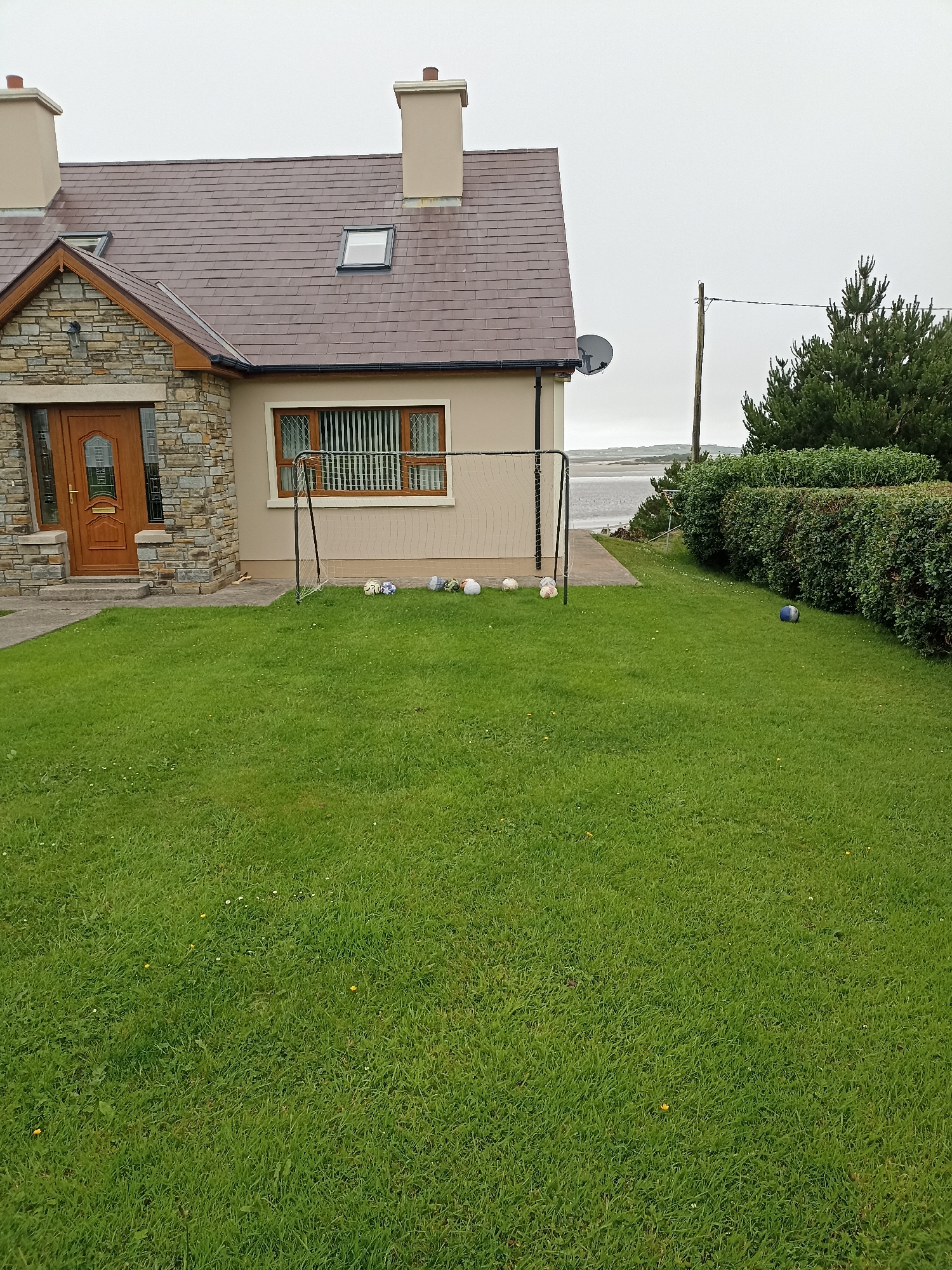 This is a medium-range exterior shot of a suburban house in Annagry West, Ireland, on an overcast day. The house, a two-story structure with a stone facade on the front left and beige siding on the rest of the exterior, features a brown tiled roof with two chimneys and two skylights. A wooden door with glass panels is visible on the front left.
In front of the house, a well-maintained green lawn stretches out, dotted with small white and yellow flowers. A small soccer goal with a net is set up on the lawn. Several soccer balls are scattered around the goal, including one blue and white ball on the far right of the lawn. Behind the house, a dense green hedge lines the property, and beyond that, a body of water is visible, likely the Atlantic Ocean, with a distant shoreline under a gray sky. A single utility pole with wires stands to the right of the house.
