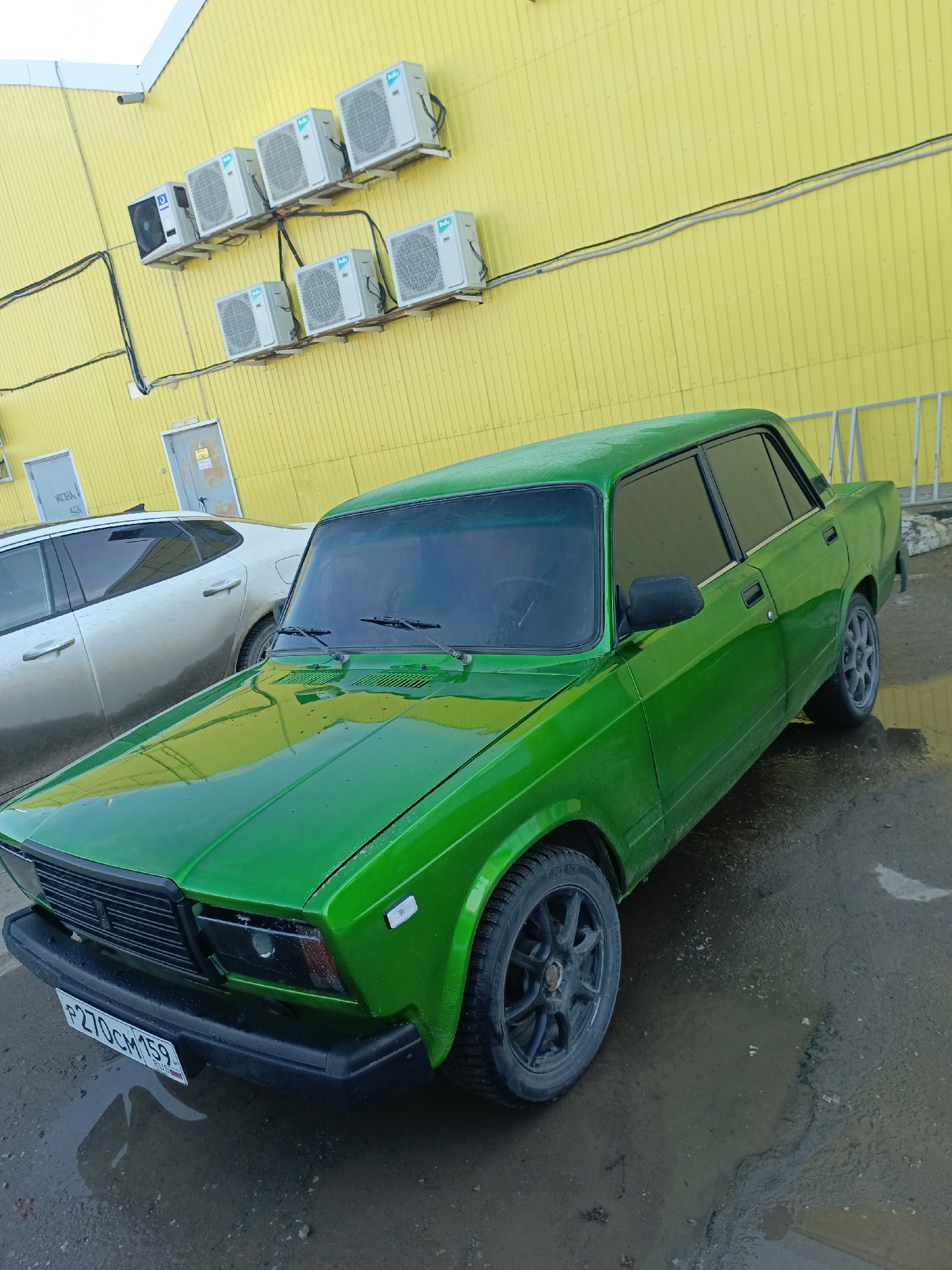 The image depicts a bright green VAZ-2107 sedan parked on a wet surface. The car is positioned diagonally, facing slightly towards the left. In the background, a yellow corrugated metal wall is visible, adorned with several air conditioning units. A silver car is partially visible to the left of the green car. The license plate of the green car reads "P270CM159," which is a Russian license plate. The "159" region code indicates that the vehicle is registered in the Perm Krai region of Russia. Chaikovsky is located in the Perm Krai region of Russia.