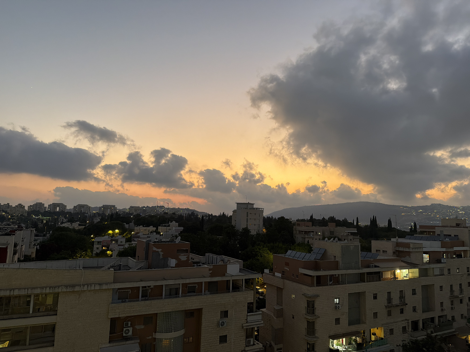 This is a horizontal, eye-level, medium shot of a cityscape at dusk in Karmiel, Israel. The sky is a gradient of soft orange and gray, with a few dark, heavy clouds dominating the upper right portion of the frame. The sun appears to be setting or has just set, casting a warm glow on the horizon.
In the foreground, multiple buildings with beige and brown facades are visible. These buildings have numerous windows, some of which are illuminated, suggesting occupied apartments. Balconies with railings are also apparent on several levels. Some of the buildings have flat roofs with visible air conditioning units and solar panels.
In the middle ground, more buildings and dense trees are scattered throughout the urban landscape. The lights of some buildings and streetlights are visible, adding to the twilight atmosphere.
The background features a range of hills and mountains, silhouetted against the fading light. The distant city lights are faintly visible on the hillsides, suggesting a wider urban sprawl. The overall scene depicts a residential area of Karmiel during the transitional period between day and night. No people or specific actions are discernable.