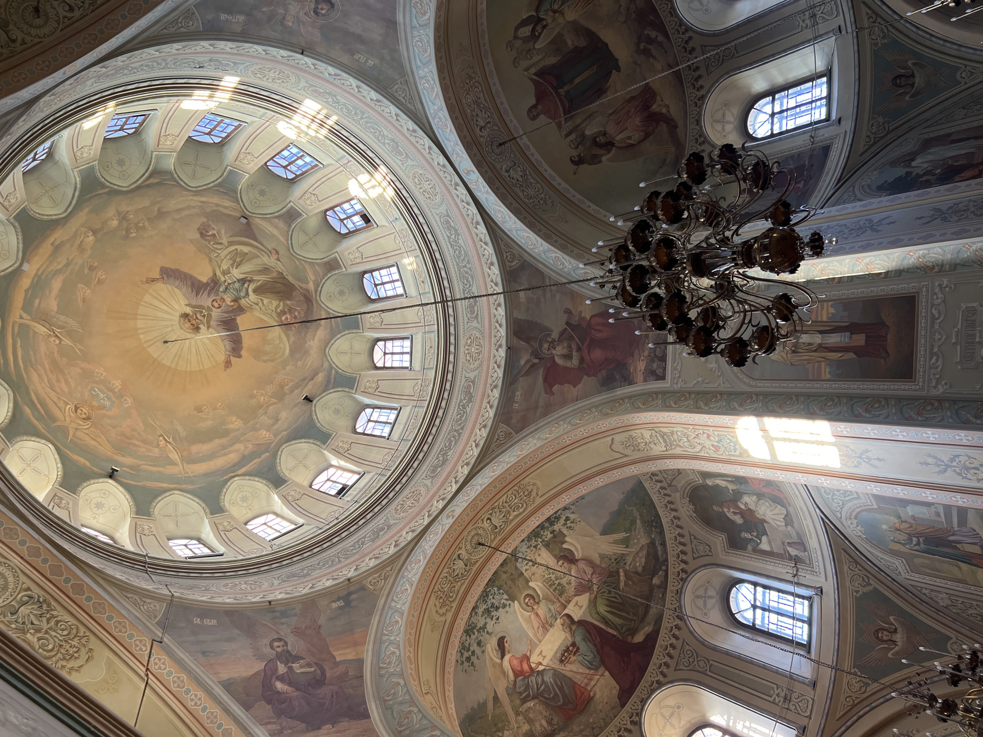 The image shows the interior of a church in Sviyazhsk, Russia, featuring painted ceilings. Several domes are visible, each adorned with religious murals depicting figures and scenes. Windows provide natural light, creating bright spots on the surfaces. A chandelier hangs from the ceiling, adding to the architectural detail.