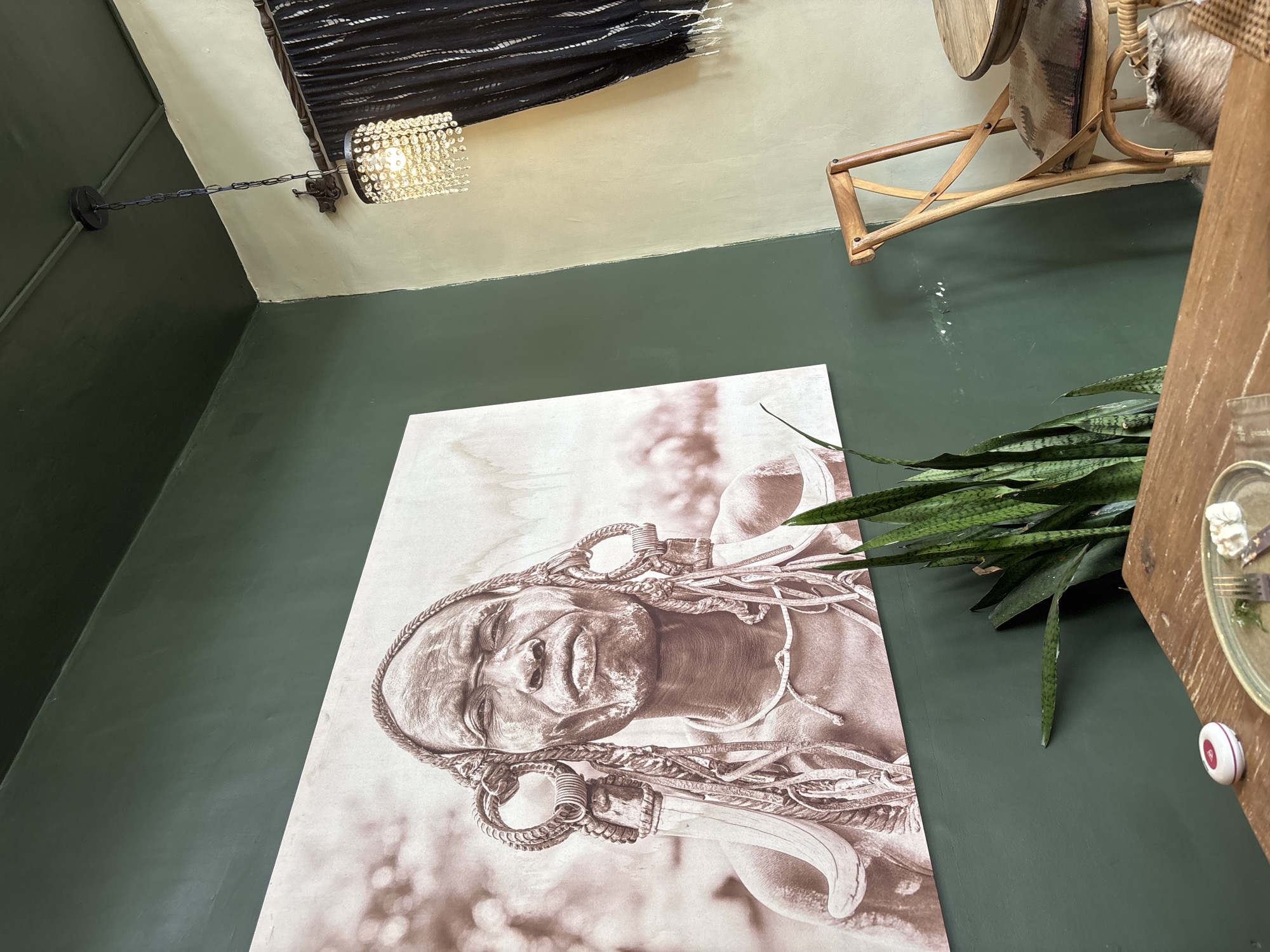 This image presents an interior scene, likely within a cafe, restaurant, or lounge, characterized by artistic decor and warm lighting. The most prominent feature is a large, sepia-toned wall art piece depicting a close-up portrait of a person, possibly indigenous, with a serene or contemplative expression. The individual's head is adorned with elaborate, intricate hairstyles featuring braids or rope-like elements, alongside decorative rings and a large, curved white object resembling a tusk or horn. The artwork rests against a dark green wall, which transitions to a lighter, neutral-toned upper wall and ceiling.
A crystal-shaded light fixture, suspended by a chain, hangs from the ceiling, casting a warm glow. Nearby, a dark, textured fabric or woven decoration is affixed to the ceiling. In the upper right corner, a portion of a wooden, possibly rattan, chair or stool with a patterned cushion is visible.
To the right of the wall art, a potted plant with long, pointed green leaves, resembling a snake plant, adds a natural element. Below the plant, part of a wooden table or counter holds a glass plate with remnants of food and a fork, suggesting recent dining. A small, white, circular object with a red center, possibly a service call button, is also on the table. The scene evokes a culturally inspired and inviting atmosphere. No discernible text is visible.