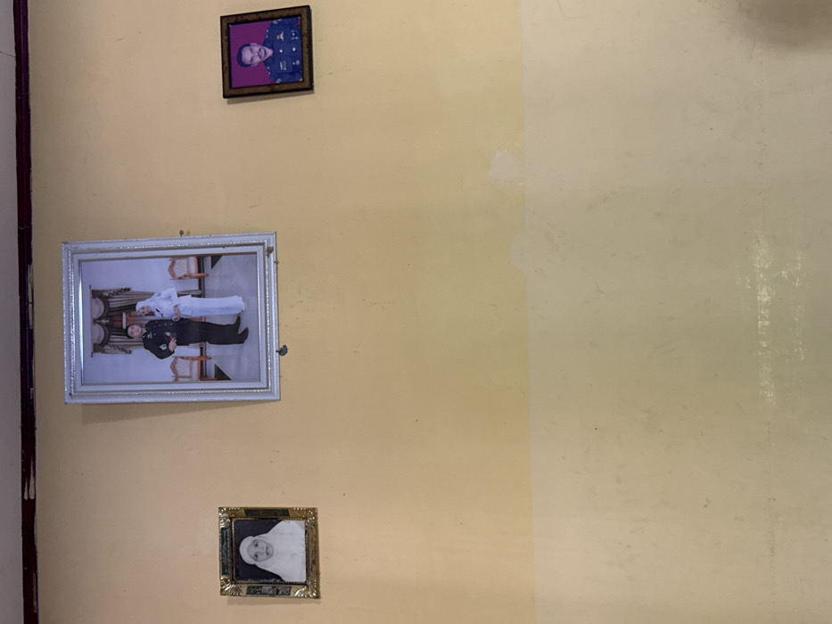 An interior wall, painted in two horizontal sections with a lighter yellow/beige tone above and an off-white tone below, displays three framed photographs. The paint shows signs of wear, scuff marks, and minor discoloration. At the top edge, a dark ceiling beam or trim is visible.

From left to right:
1.  A small, vertically oriented rectangular photograph in an ornate gold-colored frame, depicting an elderly woman with a head covering in a black-and-white or sepia-toned portrait.
2.  A larger, vertically oriented rectangular photograph in a silver-colored ornate frame, centered above the horizontal paint line. It features a full-body studio portrait of a man in a dark uniform and a woman in a white uniform dress and white hijab, standing side-by-side. The background includes two ornate chairs and curtains.
3.  A small, vertically oriented rectangular photograph in a simple dark brown frame, depicting a man from the chest up in a dark blue military or official uniform with visible insignia.

The specific location within the city of Pante, Indonesia, is not identifiable from the visual content of the image.