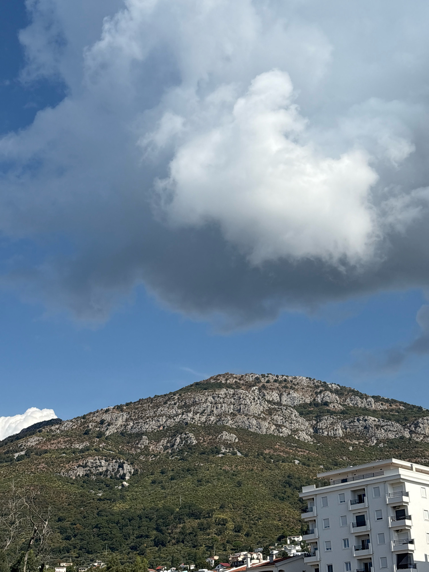 The image shows a scenic view of the city of Bar, Montenegro. In the foreground, a modern white building with balconies stands prominently. Behind it, a verdant mountainside covered in lush green vegetation rises towards the sky. The rocky peaks of the mountain are visible, adding texture and detail to the landscape. Above the mountain, a dramatic sky is filled with large, fluffy white clouds contrasted against a bright blue backdrop. A large, darker grey cloud formation looms over the scene, suggesting a change in weather. The overall impression is one of natural beauty combined with urban development, characteristic of a coastal city like Bar.