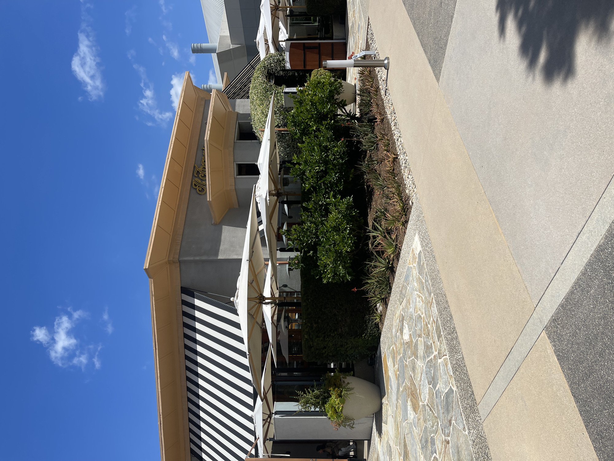 The image shows a partial view of a building with a black and white striped awning in Irvine, California, USA. The building appears to be a restaurant or business, possibly "Il Fornaio" based on visible signage. There are outdoor patio umbrellas and greenery along the walkway adjacent to the building. The walkway is paved with a combination of materials, including flagstone and concrete. The sky is blue with scattered clouds. The photo appears to be taken during daylight hours.