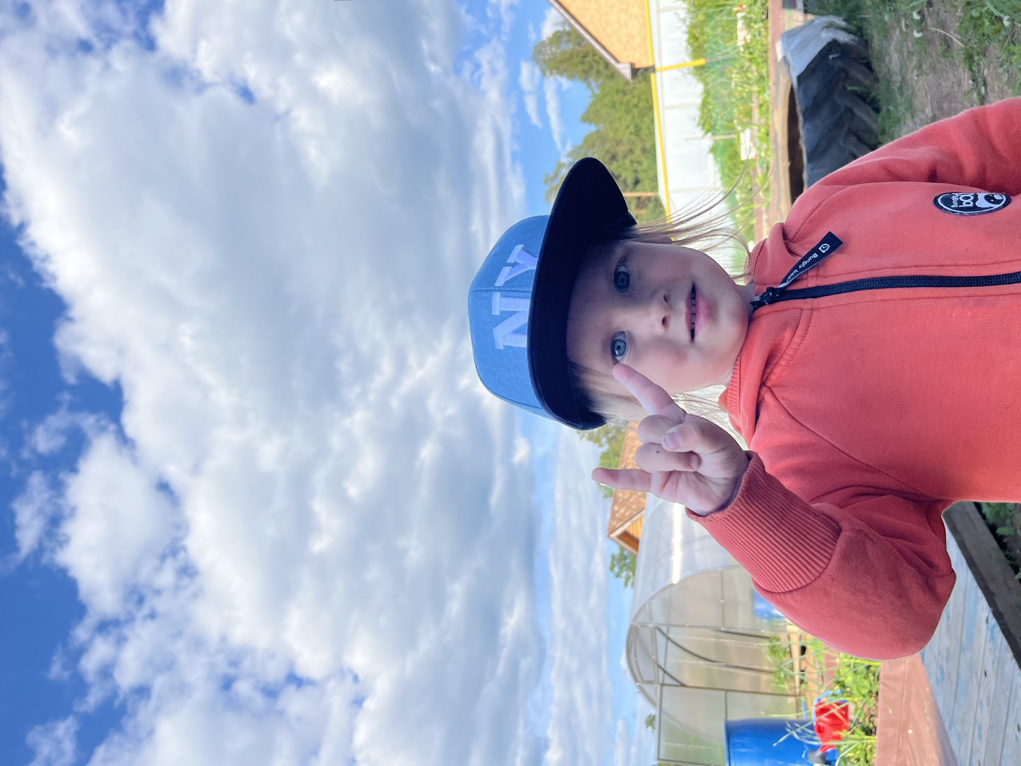 A young child, approximately 3-6 years old, is depicted in a full-frame outdoor portrait in the city of Chaikovsky, Russia. The child, with light hair partially visible, has blue eyes and a slight smile. They are wearing a light blue baseball cap with a dark blue brim, featuring "NY" in white letters on the front, and an orange/coral-colored hooded zip-up sweatshirt with ribbed cuffs. A black zipper pull with "Bungy Boo" text and a round black patch with a white panda logo are visible on the sweatshirt. The child's right hand is raised, performing a "rock and roll" gesture with the index and pinky fingers extended and the thumb holding down the middle and ring fingers. A small dark spot is visible on the back of the hand.
The background reveals a rural or semi-rural environment under a clear blue sky dotted with numerous white cumulus clouds. To the left, a large, clear tunnel-style greenhouse with a metal frame is prominent. Further left, a blue barrel and a red object are partially visible. Behind the greenhouse, a wooden cabin-like structure with a brown roof is discernible. To the right, another building with a light brown facade and dark roof is visible. A low white fence and a yellow vertical pole run across the mid-ground. A large, dark tire is laid on the ground in the mid-right. Green vegetation, indicative of garden beds, and scattered trees are visible throughout the background. The child stands on a reddish-brown surface in the immediate foreground.