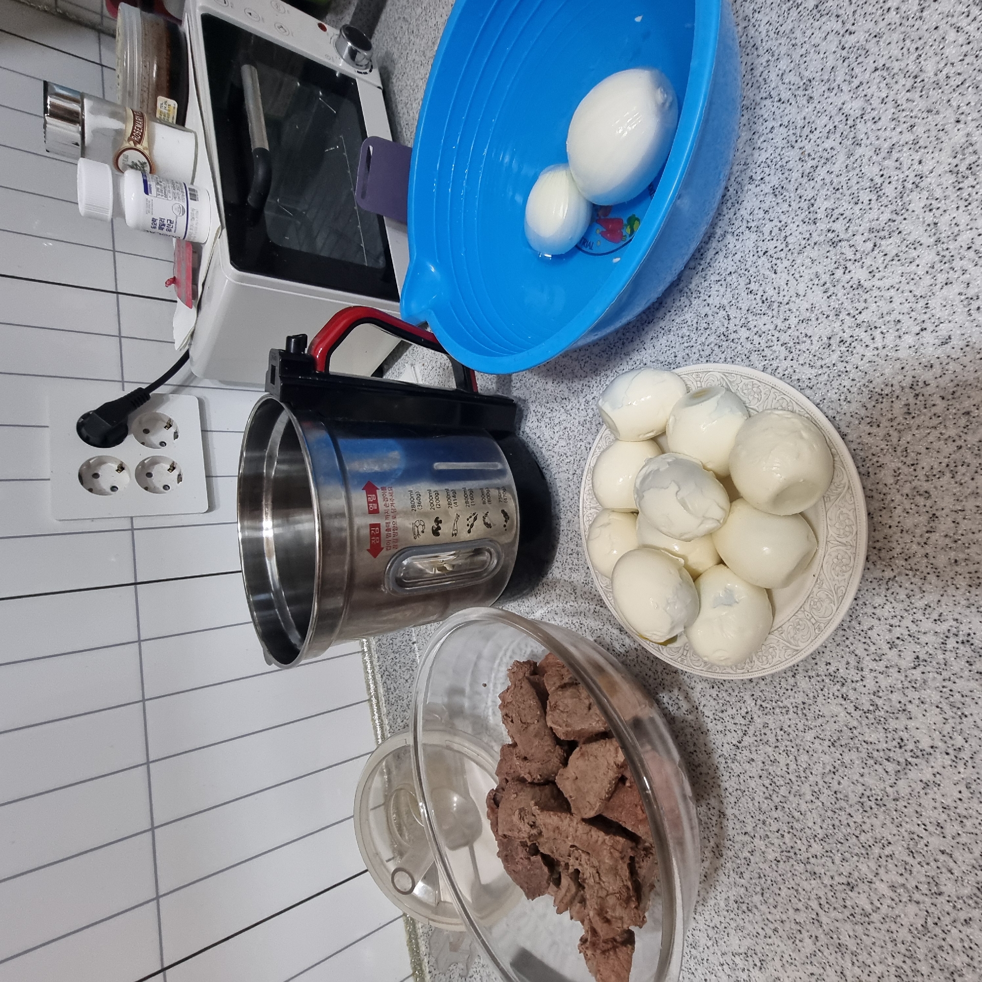 In Dunnae-myeon, South Korea, a countertop scene unfolds with food preparation items. In the foreground, a white patterned plate holds several peeled hard-boiled eggs. To the left, a clear glass bowl contains numerous chunks of cooked meat. Behind the eggs, a stainless steel multi-cooker with red accents is present, displaying measurement markings in Korean. To the right, a bright blue plastic basin holds two more peeled hard-boiled eggs. In the background, a white microwave oven is visible, with a bottle of "Rosemary" seasoning and another small bottle on top. The area is tiled with white squares and grey grout, suggesting a kitchen environment.