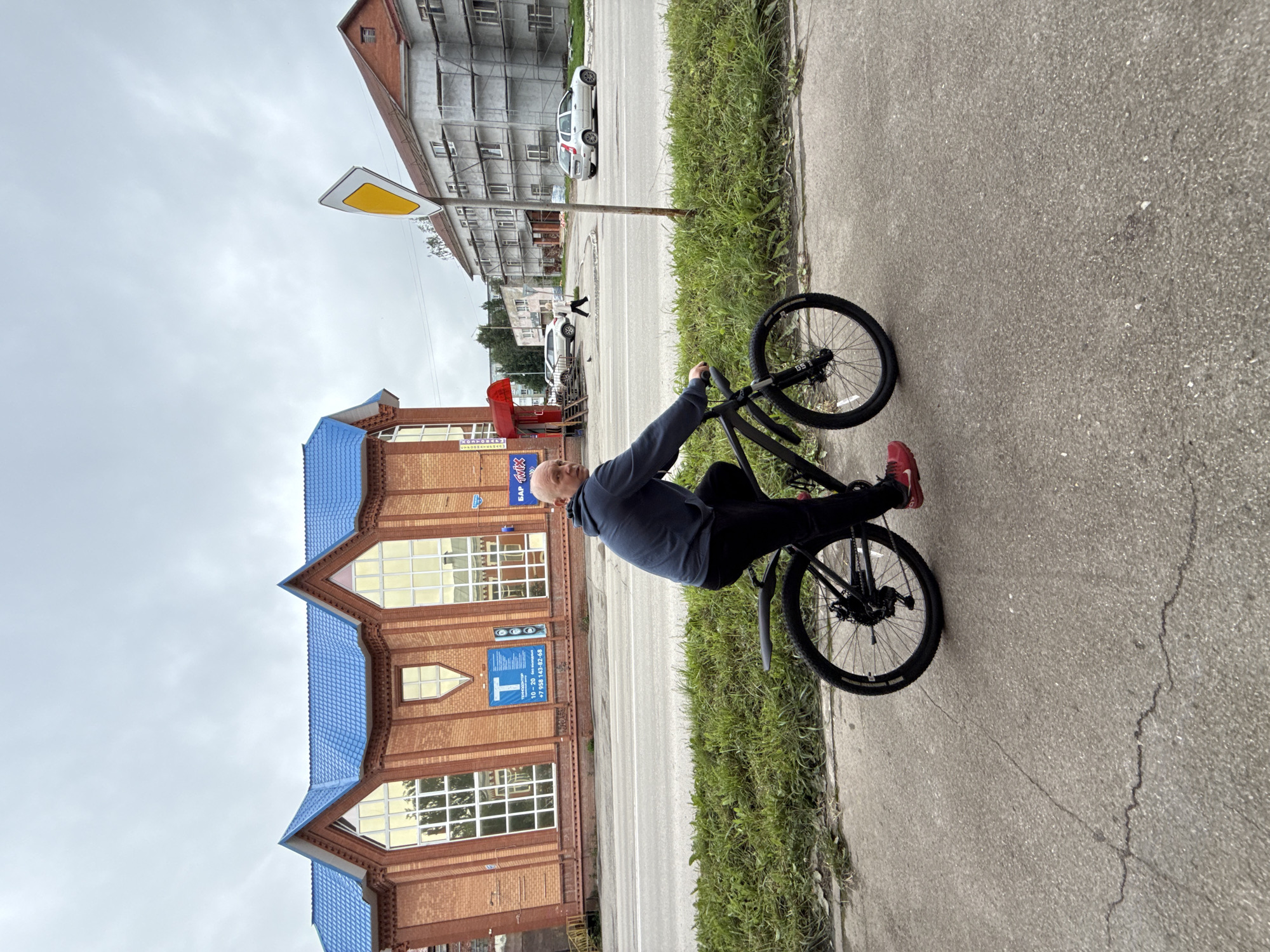 A bald man in a dark blue hoodie and black pants rides a black bicycle with red shoes on a paved sidewalk in front of a brick building with a blue roof. The building has large windows and a blue sign with Russian text. A yellow diamond-shaped road sign is visible to the right of the building. The street in front of the building has a few cars and people walking. The sky is overcast. The scene is likely located in Lysva, Russia, given the presence of Cyrillic text on the building's sign.