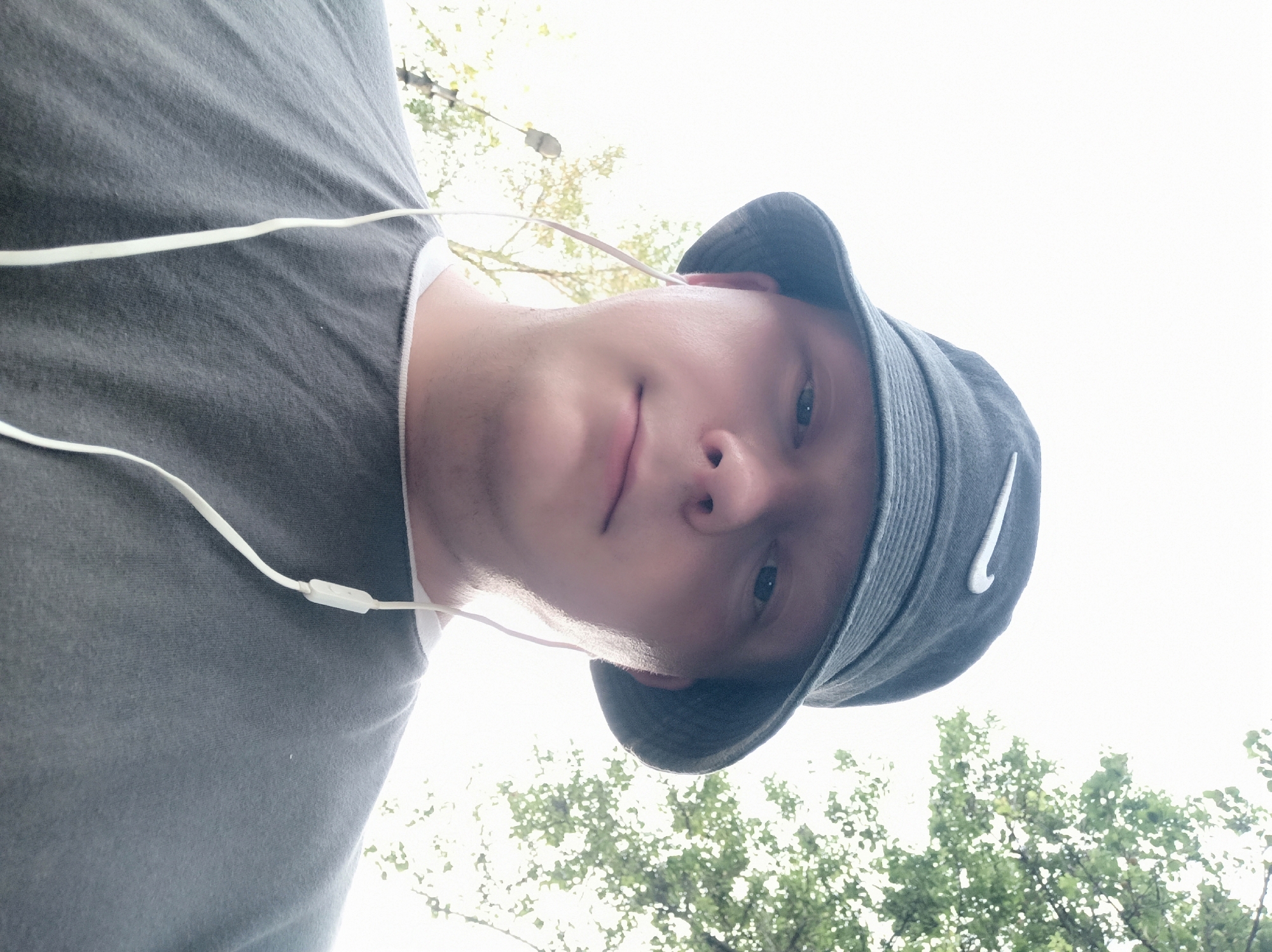 A waist-up, low-angle outdoor selfie shows a person wearing a dark bucket hat with a white Nike logo and a gray t-shirt with white trim around the neckline. White earbuds are plugged into the person's ears, with the white cord running down their shirt. The background is a bright, overcast sky with green trees visible in the upper right corner and a faint silhouette of a lamp post on the left. The person's face is looking directly at the camera.