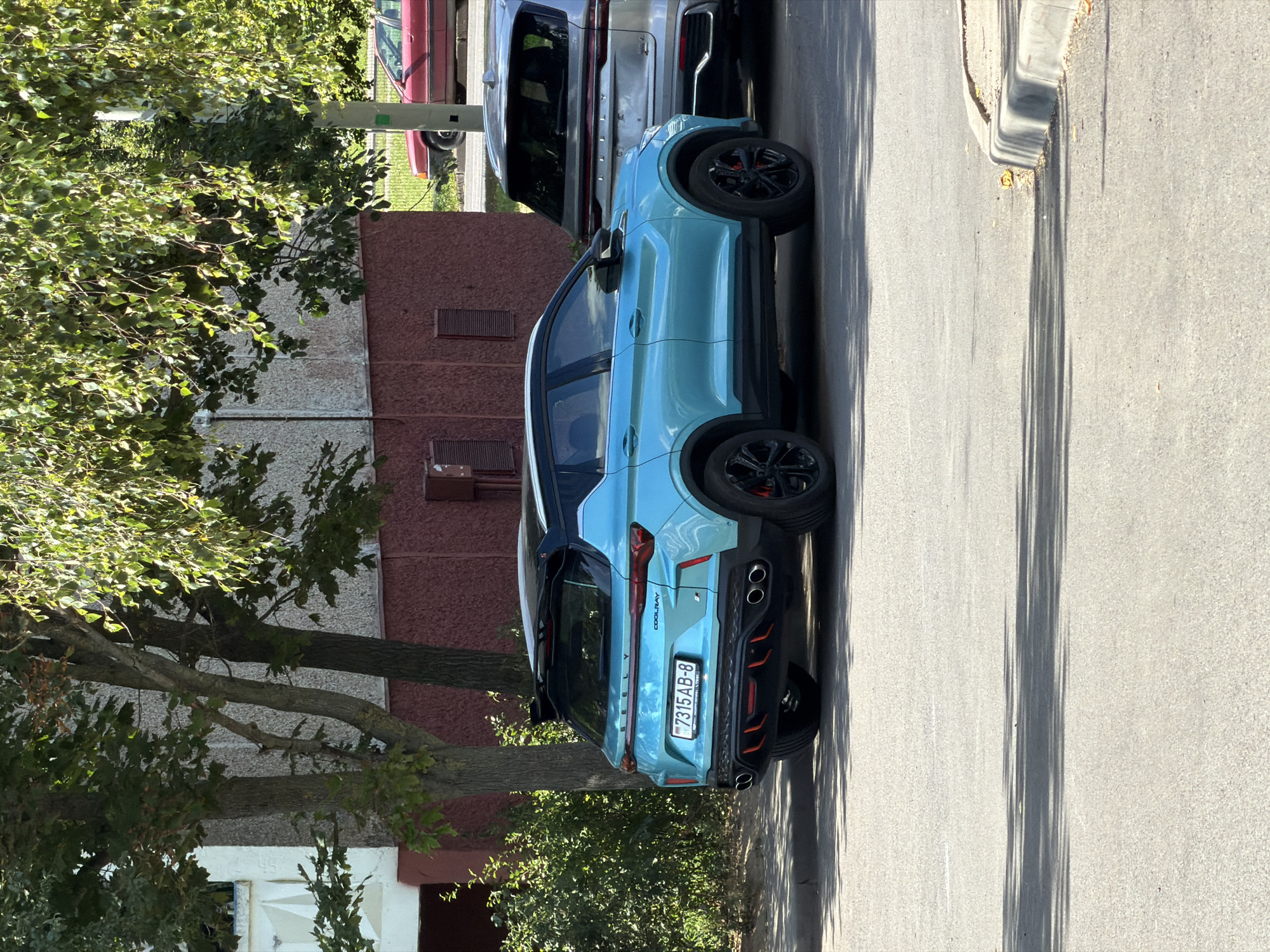 A bright blue Geely Coolray SUV is parked on a city street in Minsk, Belarus. The car is positioned on the right side of the frame, facing away from the viewer and angled slightly to the left. It features black accents, including the roof, spoiler, and wheels with red brake calipers. The license plate is visible and reads "7315 AB-8".
Behind the Geely, a silver SUV is parked, partially obscured. To the left of the Geely, a building with red and beige siding is visible, with trees and greenery in the foreground. Further in the background, a red sedan is parked on the other side of the street. The pavement is grey asphalt, and a concrete curb is visible in the lower right corner. The scene is illuminated by sunlight, casting shadows from the trees onto the street.