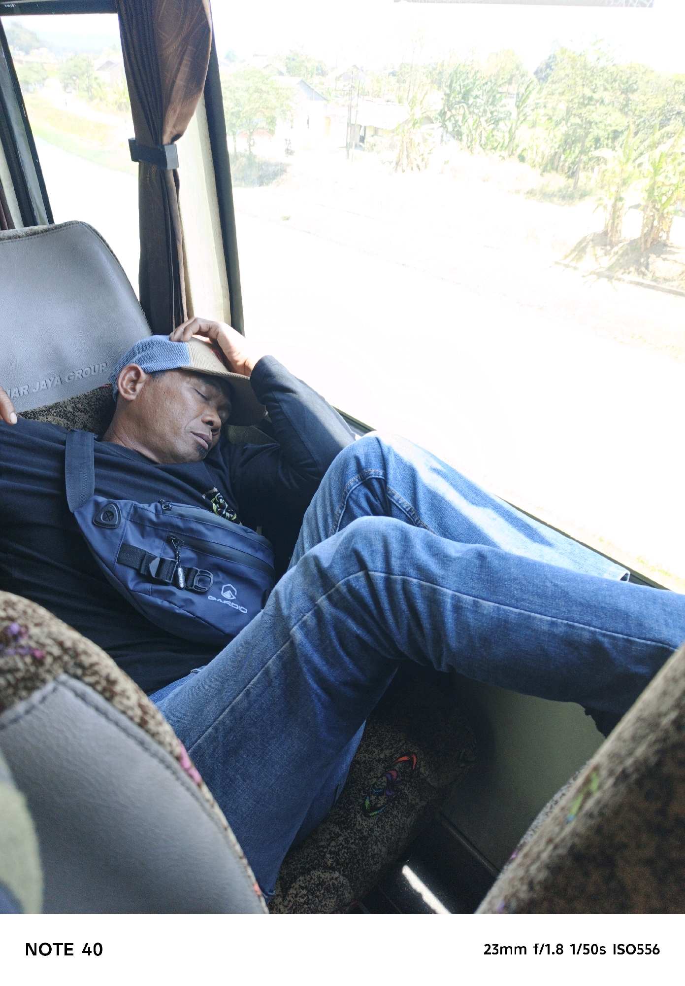 A man is sleeping on a bus with his head resting on his hand, which is wearing a cap. He is wearing a black t-shirt, blue jeans, and a blue fanny pack across his chest. The bus is moving through a rural area with trees and houses visible outside the window. The sun is shining brightly, casting shadows on the man's face and legs. The seats are covered in a patterned fabric. The image is taken from a low angle, looking up at the man. The text at the bottom of the image indicates that it was taken with a 23mm lens at f/1.8, 1/50s shutter speed, and ISO 556.
