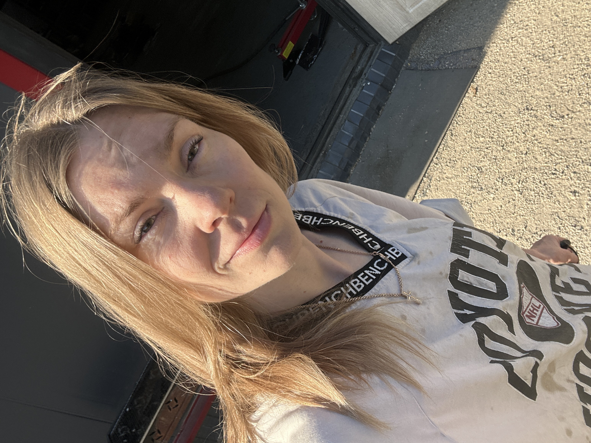 A woman with blonde hair is taking a selfie. She is wearing a light-colored t-shirt with a black logo that includes "BENCH" written in white letters and an NHL logo. She has a delicate gold chain with a cross pendant around her neck. Her hair is loose and falls around her face.
The scene appears to be outdoors, with sunlight casting strong shadows. The ground is covered in gravel, and there's a paved area with some brickwork visible. In the background, a dark structure, possibly a garage door or wall, is visible with a red element. Part of what looks like a car jack is also present.
The lighting suggests it is daytime, likely late afternoon due to the warm, angled light. The woman's expression is relaxed and she is looking towards the camera. The overall atmosphere is casual and possibly related to some outdoor activity or maintenance work given the setting and the slightly stained t-shirt. The image is cropped closely on the woman's face and upper body, with the background providing context for the outdoor location.