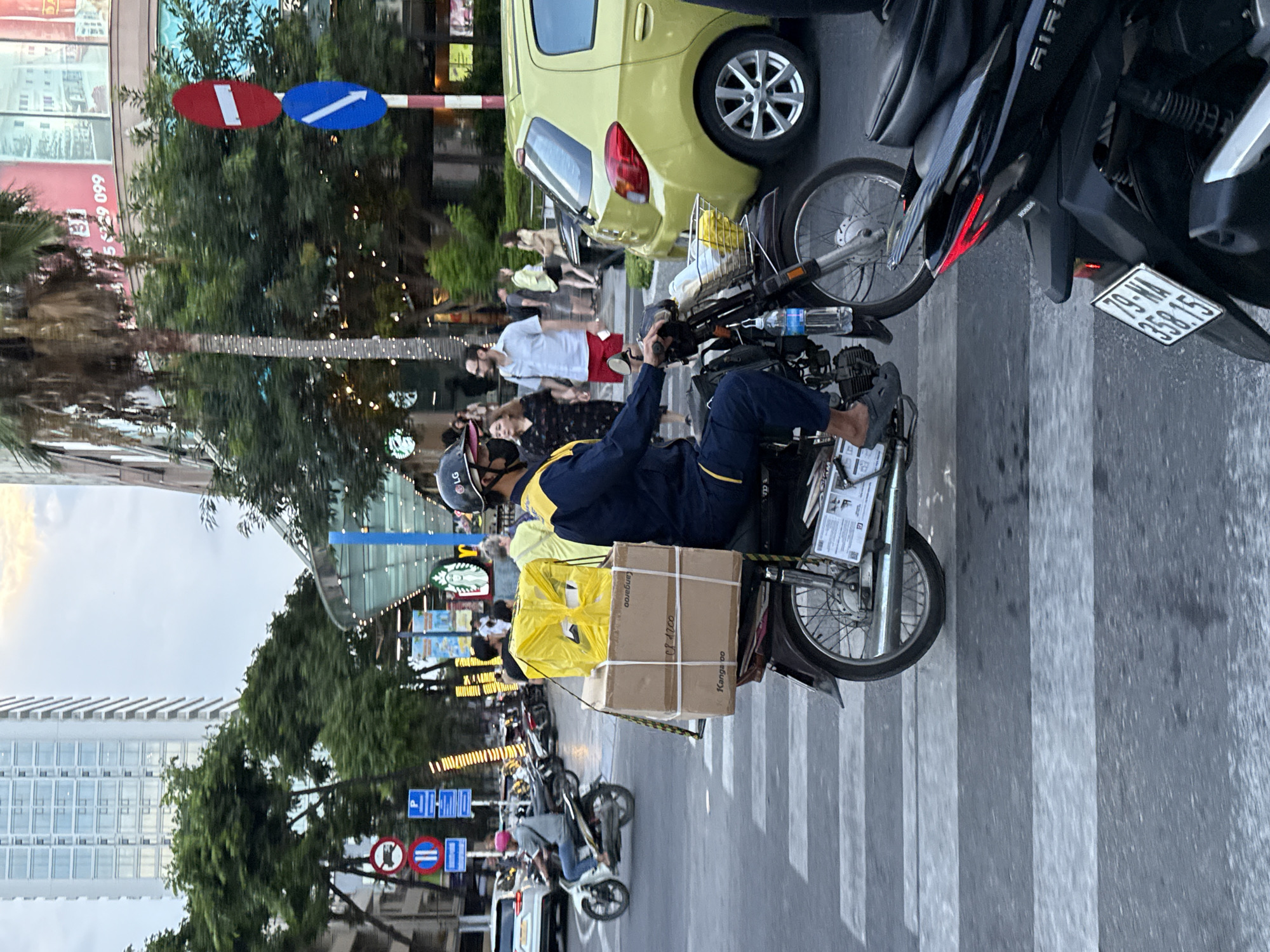 In Nha Trang Ward, Vietnam, a male delivery person wearing a blue uniform and a helmet is riding a motorcycle across a crosswalk. The motorcycle is carrying two large cardboard boxes on the back, one stacked on top of the other, and a yellow-wrapped package on top of those. The man is wearing dark pants and dark slip-on shoes. In the background, several other motorcycles and a yellow car are visible. The scene takes place on a city street with tall buildings and trees lining the sidewalks, and streetlights are illuminated. Traffic signs are visible on the left side of the image. The sky is overcast.