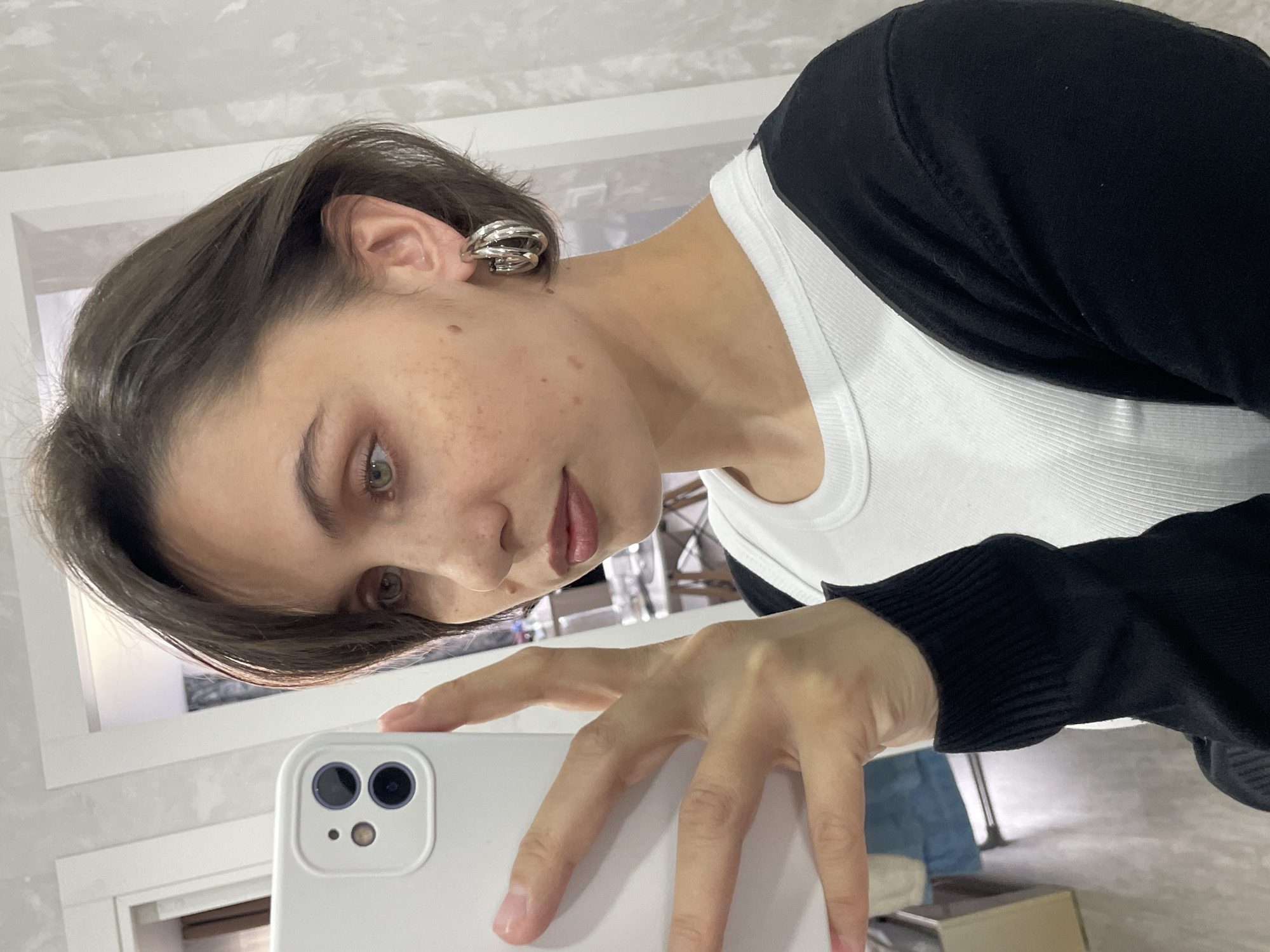 A woman is taking a selfie in a well-lit interior space. She holds a white smartphone in her right hand, partially obscuring the lower half of her face. She has short, dark hair and light-colored eyes. She is wearing a white tank top under a black cardigan. On her left ear, she wears a multiple-hoop silver earring. The background appears to be a room with white walls and a window. There is no information to suggest the location is in Izhevsk, Russia.