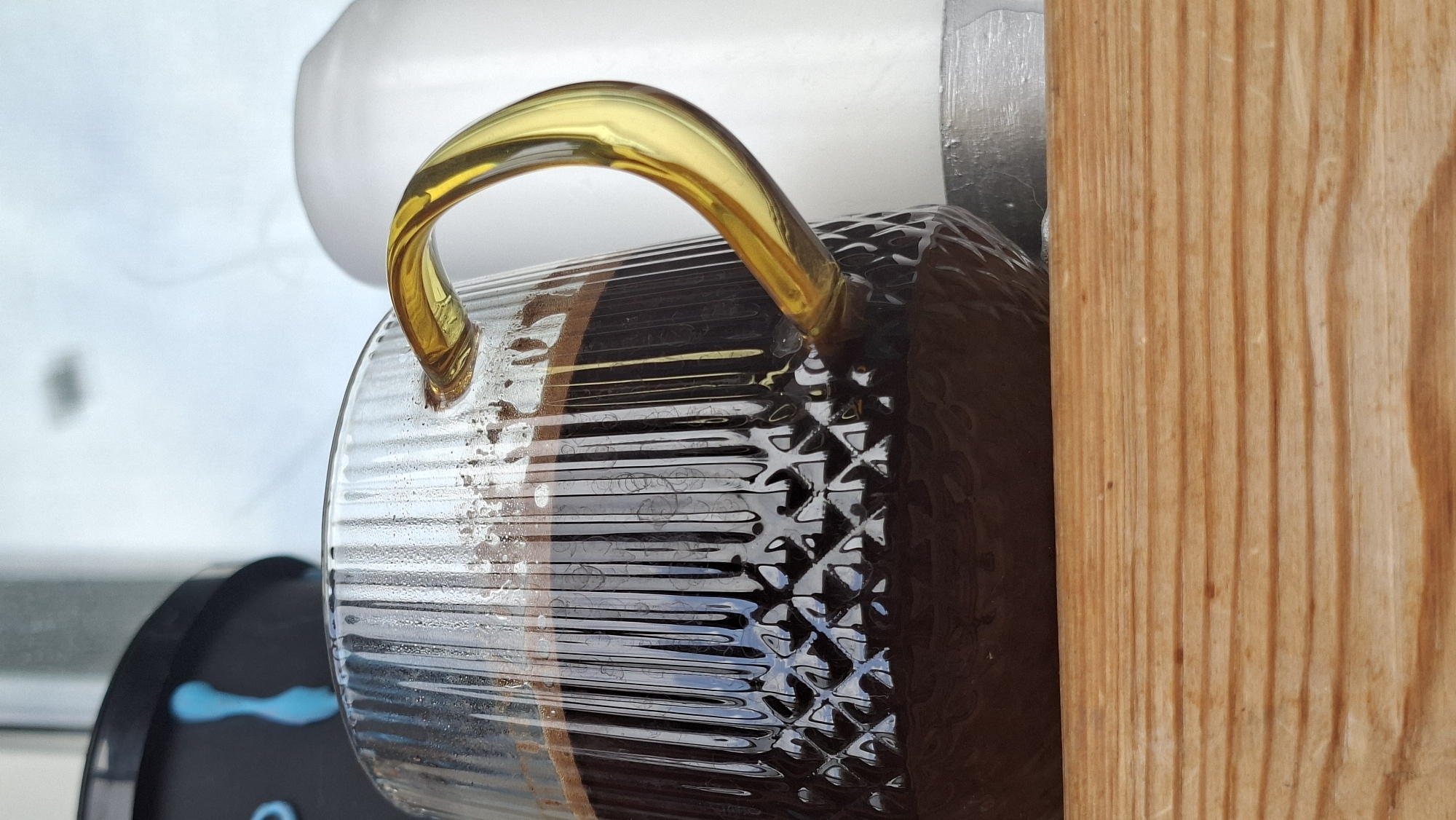 A close-up shot captures a transparent glass mug filled with dark liquid, possibly coffee, sitting on a wooden surface. The mug has vertical fluting and a textured pattern on its lower half. A translucent amber-colored handle curves gracefully from the mug. In the background, out of focus, are a black object with blue markings and a white object. The scene is likely indoors, given the lighting and the presence of objects that could be home decor or kitchen items. The exact location within Забалоцкі сельскі Савет, Belarus cannot be determined from the image alone.