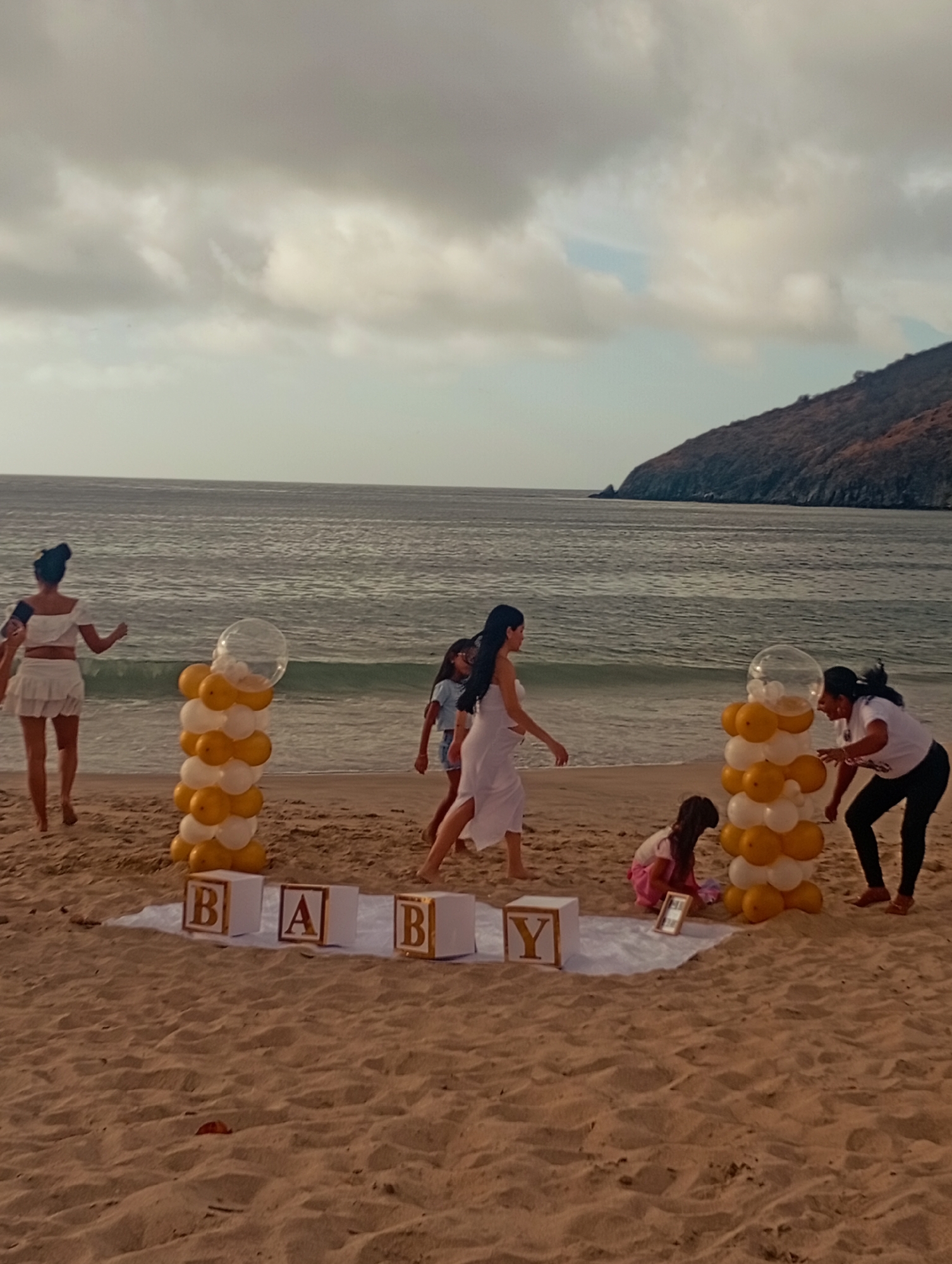 On a sandy beach in Pedro González, Venezuela, under an overcast sky, a baby shower scene unfolds. The ocean meets the shore, with a land mass visible in the distance. A group of people, including women and children, are positioned around decorations spelling "BABY" in large, white, gold-lettered blocks. Balloon pillars, arranged in white and gold, stand on either side of the letters. One woman in white stands apart, while others are engaged in setting up or interacting with the decorations. A blanket provides a backdrop for the scene.