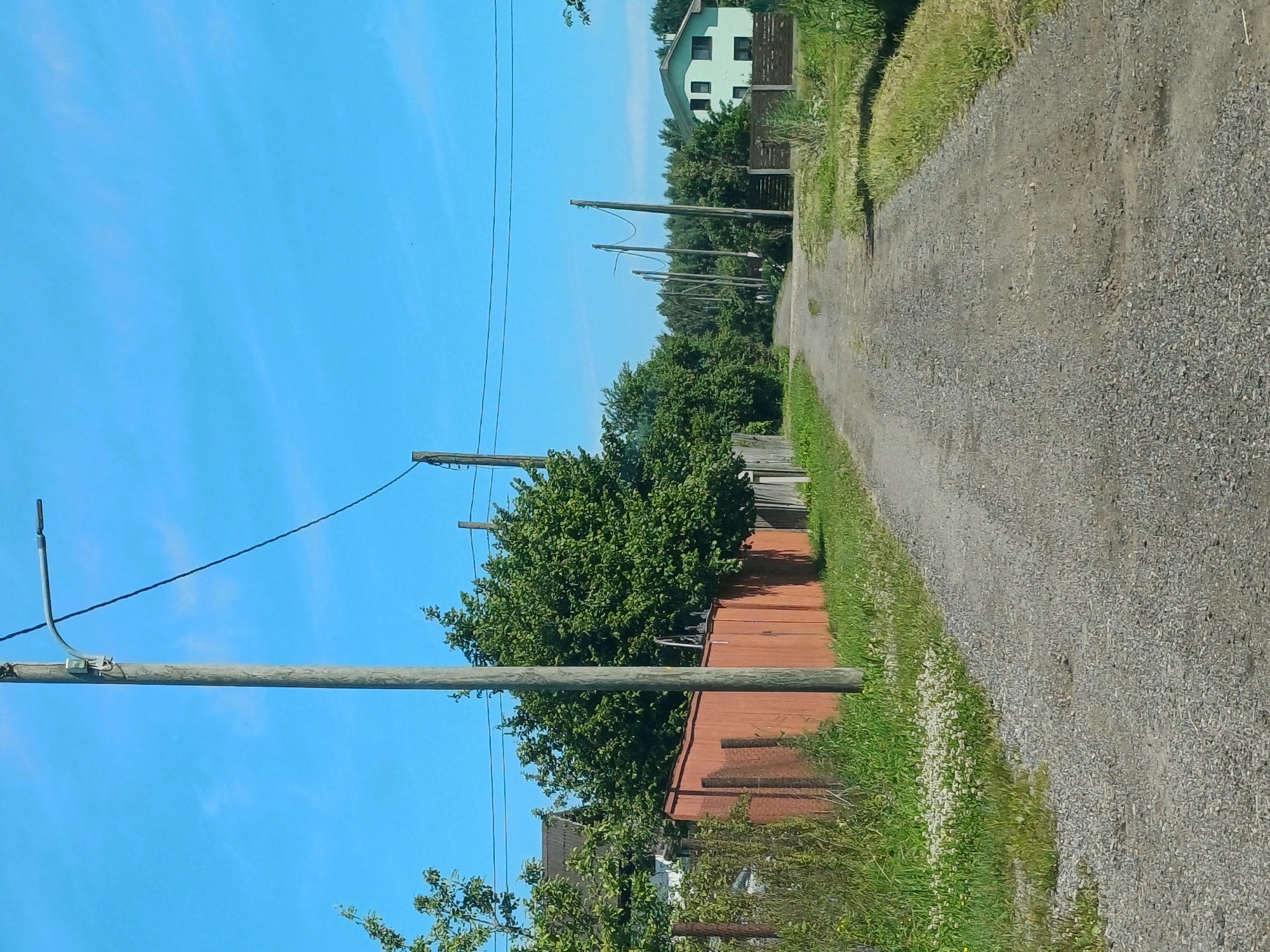 The image depicts a gravel road in a rural or suburban area. The road is lined with grass and vegetation on both sides. In the background, there are several wooden utility poles with wires strung between them. A light green house with dark windows is visible on the right side of the road. A red fence runs along the left side of the road, partially obscured by trees and bushes. The sky is clear and blue with a few wispy clouds. The overall impression is that of a quiet, undeveloped area. It's not possible to determine the specific location within Riga, Latvia based solely on this image.