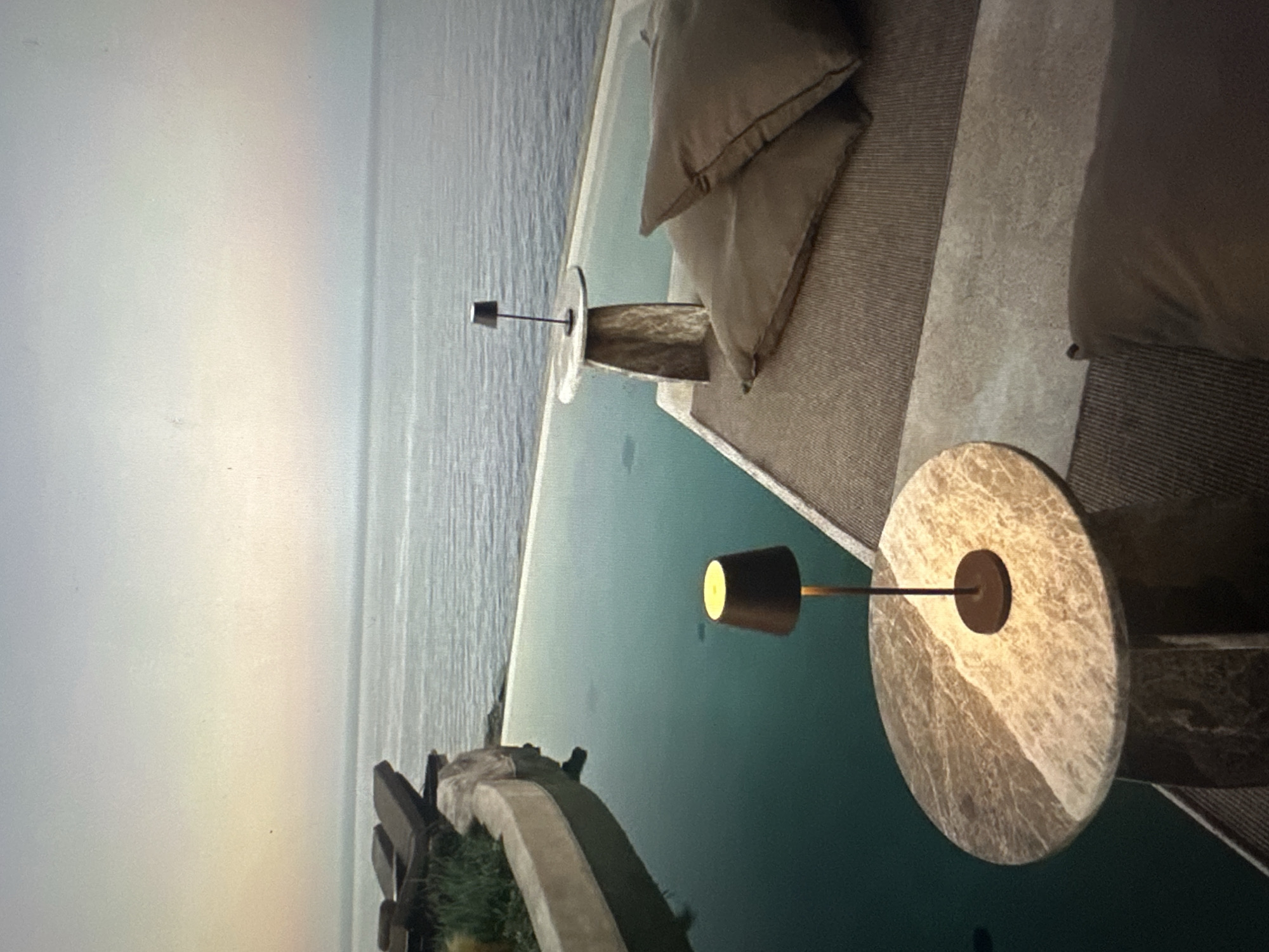 This media file captures an outdoor seating area overlooking a body of water, likely the ocean, given the gentle waves and expansive horizon. The scene is set at dusk or dawn, as indicated by the soft, diffused light illuminating the sky and the water. Two modern, low-profile lamps with metallic shades and a warm glow are placed on circular marble-topped tables. These tables have textured, possibly stone or concrete bases.
Adjacent to the tables, a lounge area is visible, featuring plush, neutral-colored cushions that suggest comfort and relaxation. The seating itself appears to be made of a textured, woven material. The swimming pool, filled with tranquil turquoise water, borders the seating area, creating an infinity edge that merges with the distant sea. In the foreground, a section of the pool's edge is visible, adorned with some greenery and a decorative stone element. The overall impression is one of luxurious serenity and an inviting space for quiet contemplation or peaceful enjoyment of the natural surroundings. No people are visible in the frame, and there is no visible text.