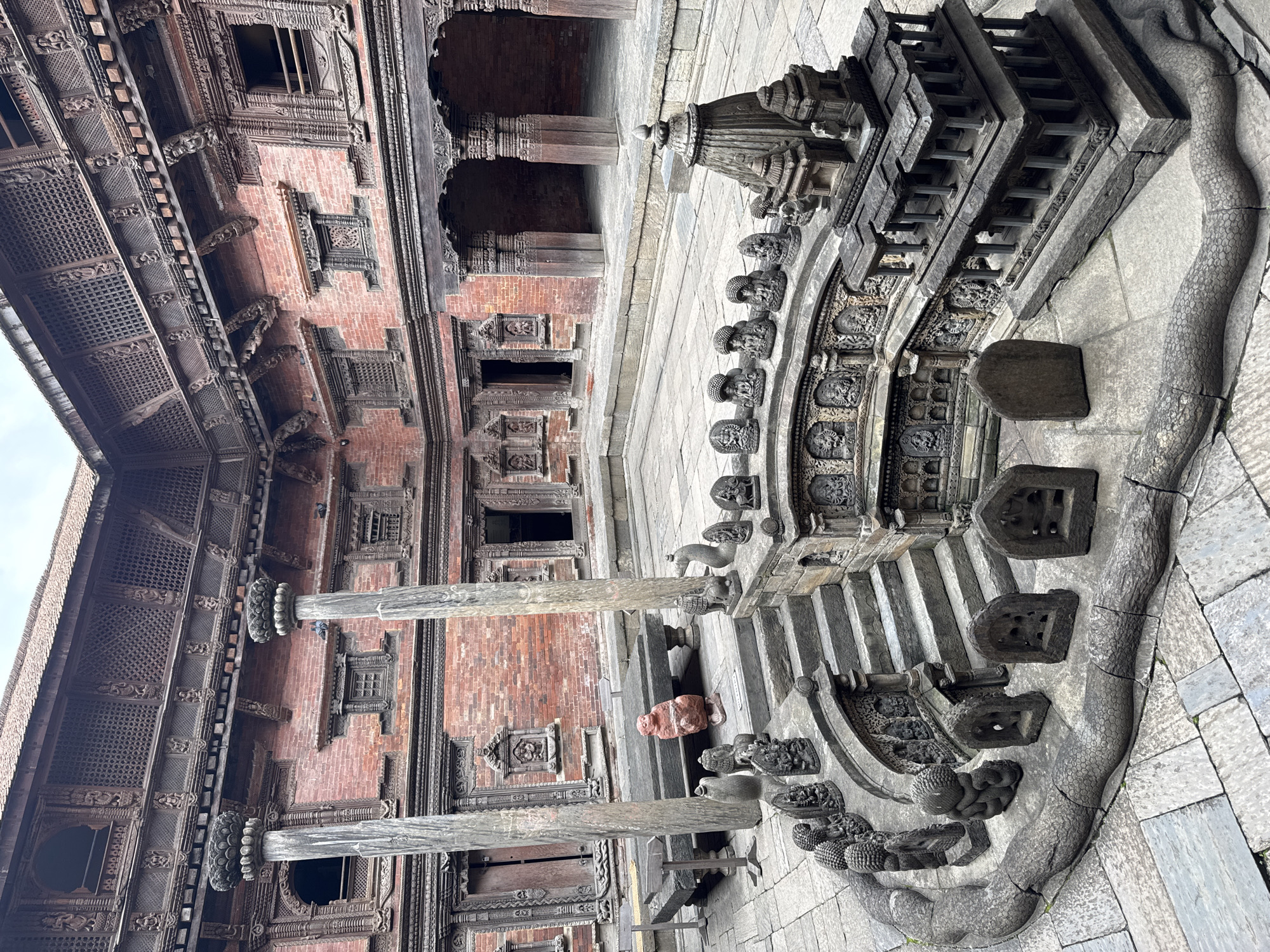 The image shows a courtyard in the Patan Durbar Square, Lalitpur, Nepal, with detailed brick and wood architecture. A stone water spout ("hiti") complex sits in the foreground, featuring sculpted figures and tiered structures. A person is sitting near the wooden columns supporting the building. The area is paved with stone.