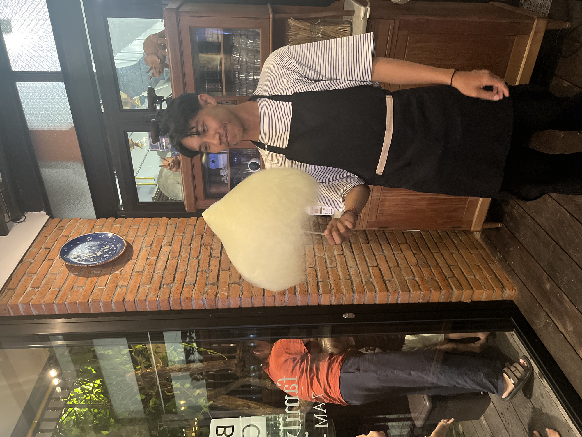 A young man, wearing a striped t-shirt and a black apron, stands indoors holding a large, pale yellow ball of cotton candy on a stick. He is looking towards the right of the frame with a slight smile. Behind him, a brick wall is visible, adorned with a blue and white decorative plate. To his left, a large glass window reflects the interior and exterior of the establishment. Through the window, a person in an orange shirt and dark pants is visible, along with other indistinct figures and foliage. The floor is made of dark wood planks. To the right of the man, a wooden cabinet filled with glassware and other items is present. The environment appears to be a cafe or restaurant in North Kuta, Indonesia.