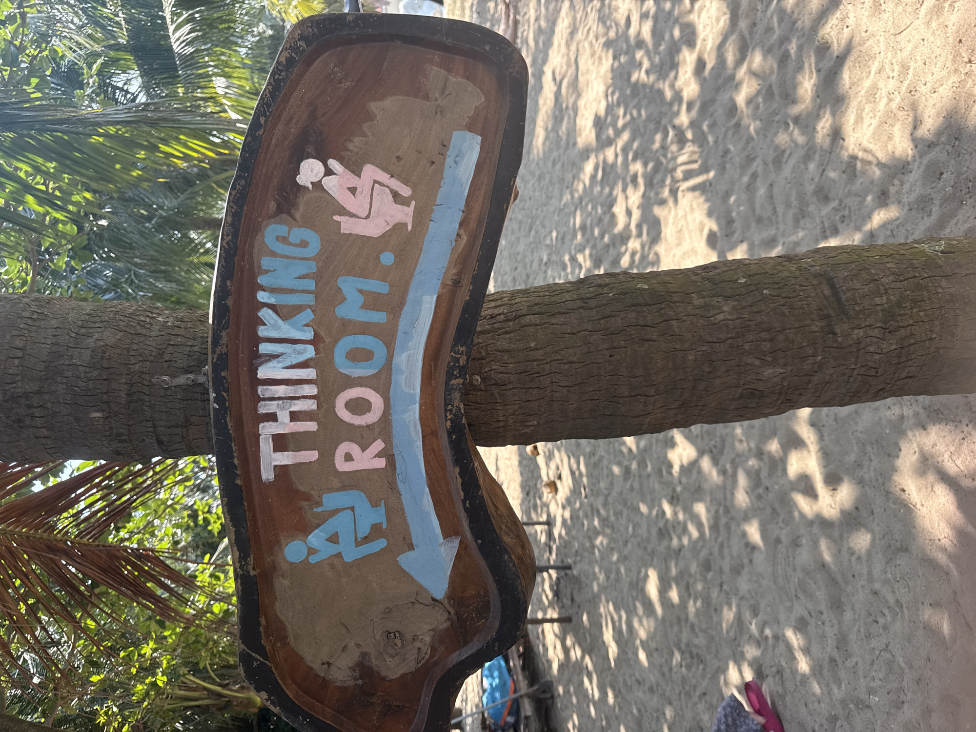 A wooden sign, irregularly shaped and stained dark brown with a black outline, is horizontally affixed to the textured, light brown trunk of a palm tree. The sign displays the text "THINKING ROOM." in white and light blue capital letters. Below the text, a light blue arrow points left. To the left of the text, a light blue stick figure sits on a toilet with its hand on its chin. To the right, a pink stick figure, distinguishable by a bun-like hairstyle, also sits on a toilet with its hand on its chin. The background above the sign consists of bright green and darker brown tropical foliage, including palm fronds, under direct sunlight. The ground below the sign is light tan sand, exhibiting various footprints and dappled shadows from overhead leaves. To the lower left, a blue object rests on a visible portion of a wooden structure. Further right on the sand, two light brown, spherical objects are present. This scene depicts a beach environment, consistent with coastal areas in Đà Nẵng, Vietnam.