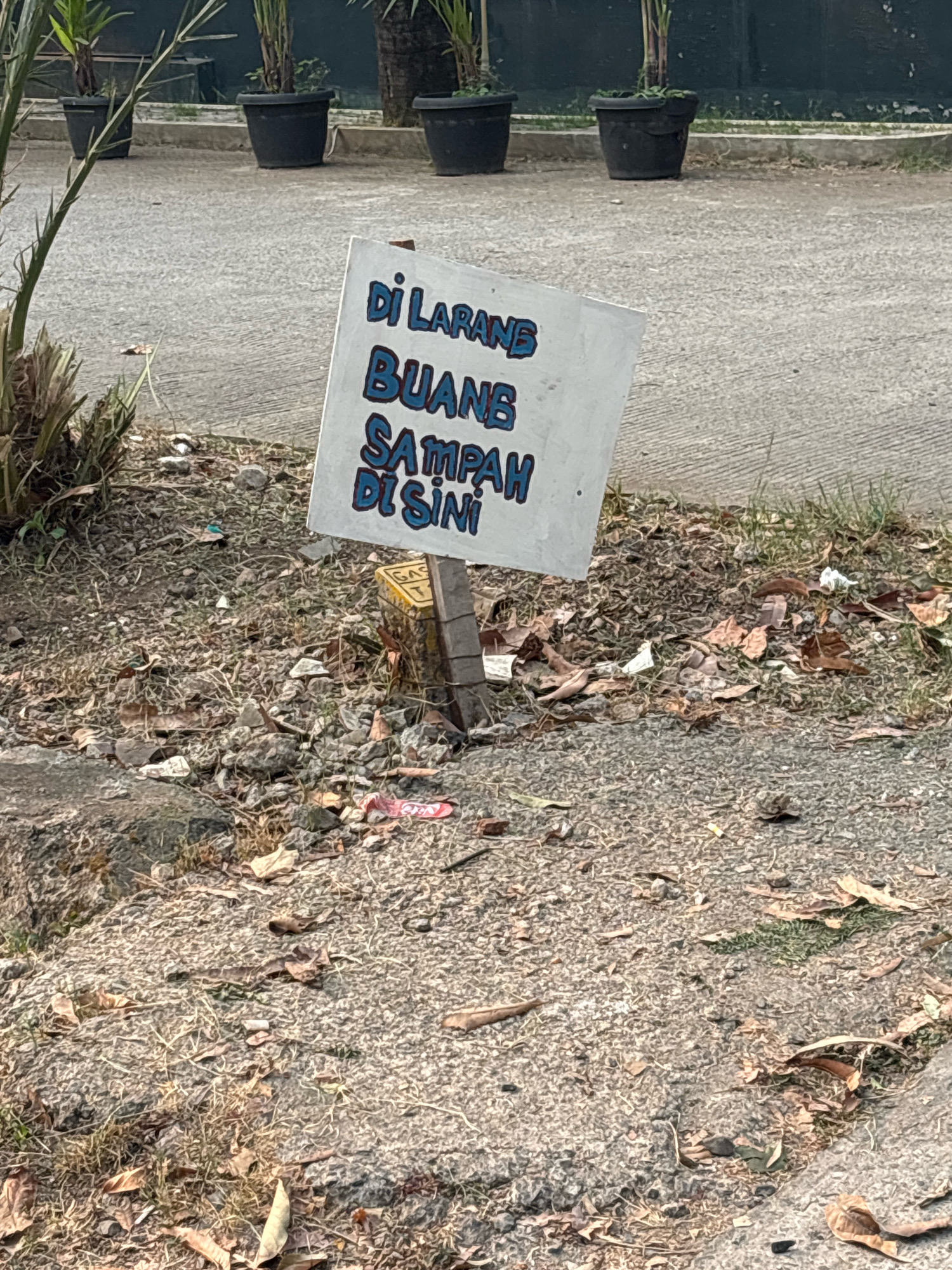 The image shows a white sign with blue text in the Indonesian language, mounted on a wooden post. The sign reads "DILARANG BUANG SAMPAH DI SINI," which translates to "DO NOT DUMP TRASH HERE." The sign is positioned outdoors, near a paved area and a patch of dry grass and fallen leaves. In the background, several black potted plants are visible, suggesting an urban or semi-urban setting, possibly in the city of Bekasi, Indonesia. The ground around the sign is littered with small pieces of trash, including wrappers and cigarette butts, despite the warning. The overall impression is one of a neglected public space.