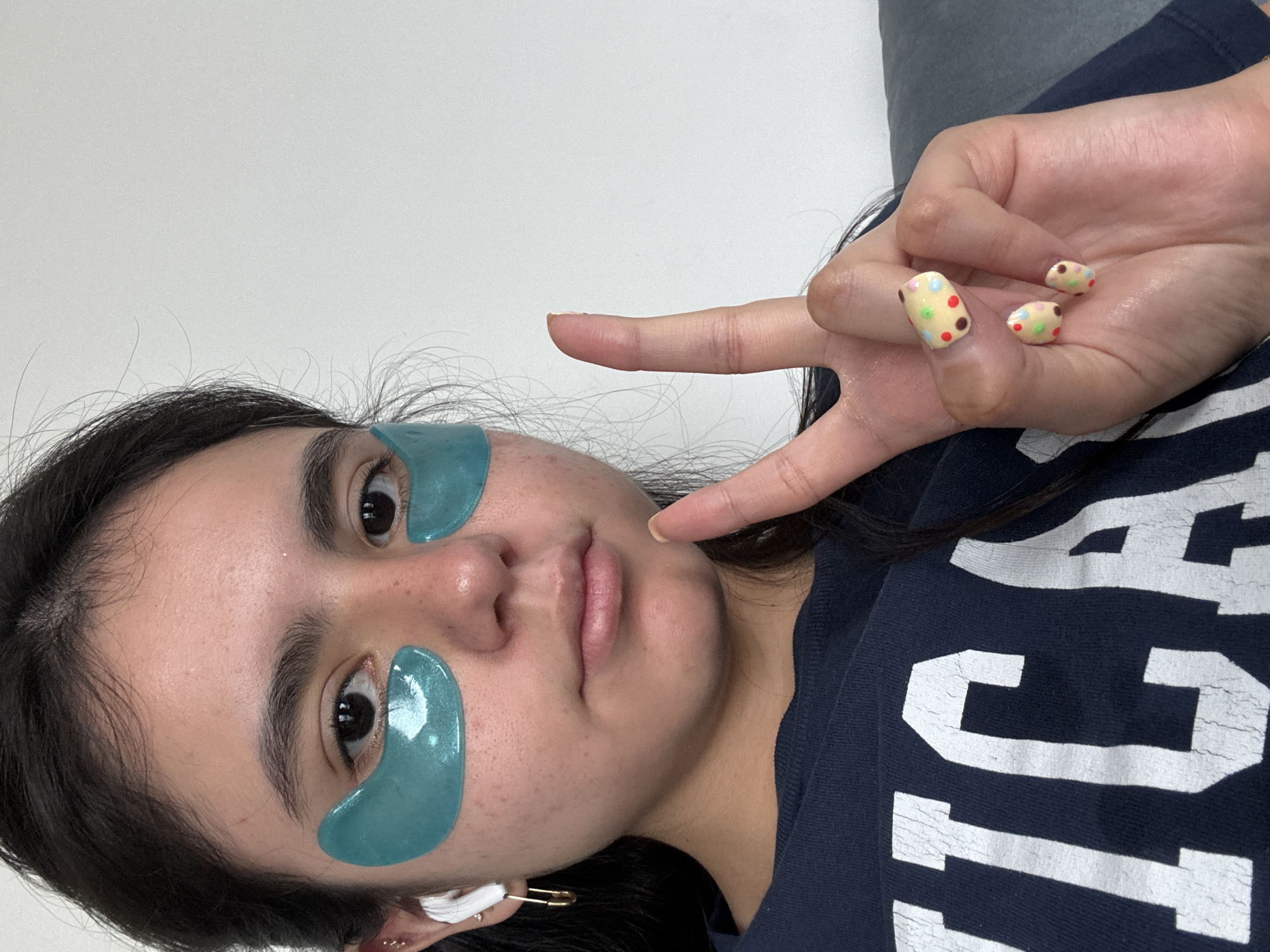 A woman is visible from the chest up, wearing blue under-eye patches. She is holding up her hand, with her index and middle finger extended in a "V" sign. Her nails are painted with a polka dot design. She is wearing a dark blue shirt with white lettering. A gold safety pin is attached to her ear, near a cotton swab. The background is a plain white wall. Without further information, it is impossible to identify the exact location as Austin, Texas, but the details are consistent with the types of clothing and accessories common in the area.