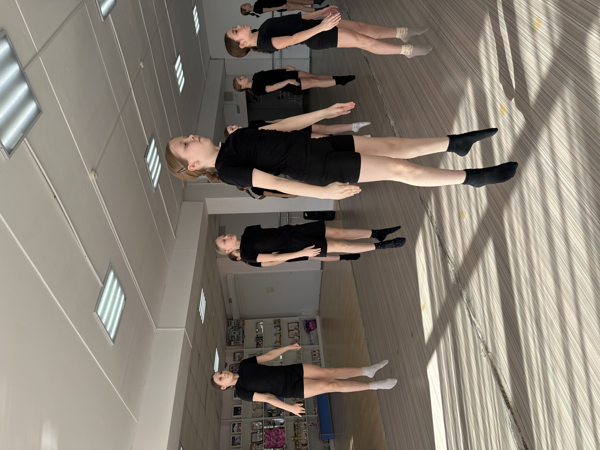 In a dance studio in Votkinsk, Russia, a group of approximately seven young females, appearing to be pre-teens, are captured mid-jump during a synchronized exercise or dance routine. All individuals are uniformly dressed in black short-sleeved shirts and black shorts. Their feet are elevated off the light-colored, striped wooden floor, which is marked by strong, elongated shadows suggesting natural light entry. The floor also shows a long, distinct seam or crack.
The participants face predominantly towards the right side of the frame, with arms slightly abducted from their torsos and legs extended. They wear various socks: white, black, and patterned, but no footwear.
The studio features white walls and a white ceiling equipped with multiple rectangular fluorescent light fixtures. On the left wall, shelving units display numerous trophies, awards, and framed photographs. The back wall includes a large, light-colored roll-down door or shutter and a dark doorway marked "ВЫХОД" (Exit). A large mirror, implied by reflections on the right side, extends the visual depth of the room. Circular smoke detectors are mounted on the ceiling.
