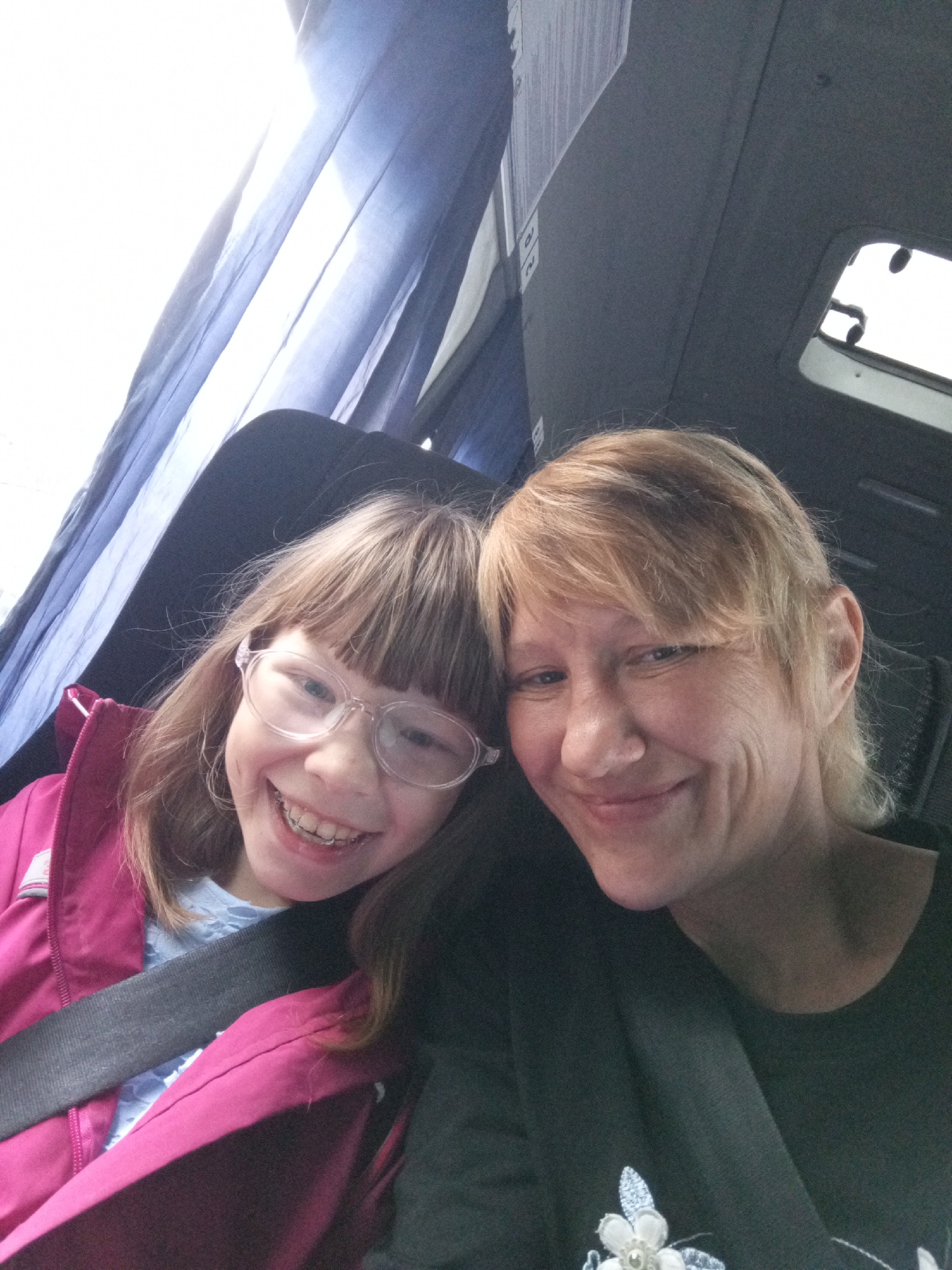 Two individuals, a child and an adult, are positioned in what appears to be the interior of a vehicle, possibly a bus or van, as suggested by the presence of seatbelts and interior paneling. The child, on the left, is wearing a bright pink jacket over a light blue shirt and has on clear-rimmed glasses. They are smiling broadly, revealing braces on their teeth. The adult, on the right, has blonde hair and is wearing a dark shirt with a floral design. Both are looking towards the camera, indicating they are taking a selfie. The background shows blue curtains and grey interior surfaces. The scene suggests a casual moment of travel. The location within Verkhneyarkeyevo, Russia, is not discernible from the visual information provided in the image.